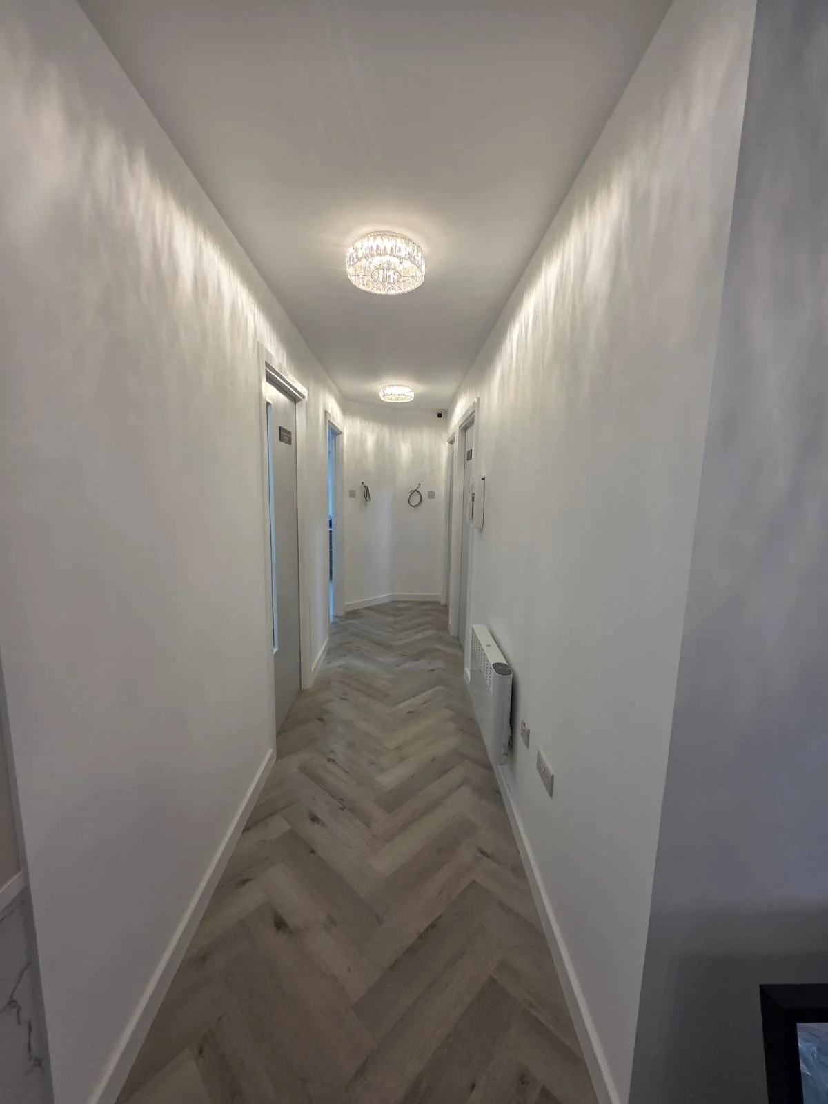 A clean, well-lit hallway with white walls and modern ceiling lights. There are three doors along the corridor, one with a sign on it. The floor has a light wood herringbone pattern, and there is a wall-mounted heater on the right side.