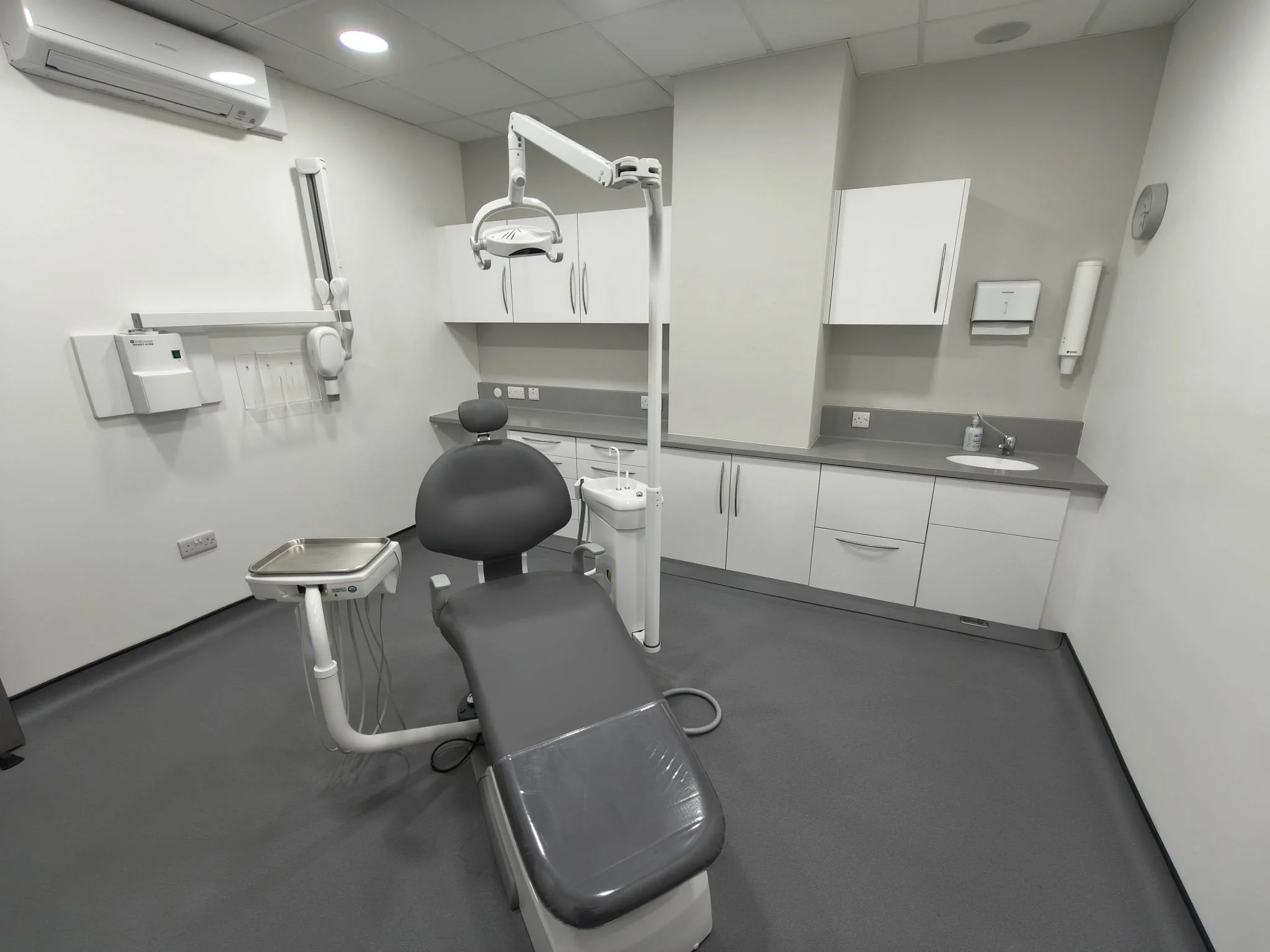 A dental examination room with a dental chair, overhead light, cabinets, and a sink.