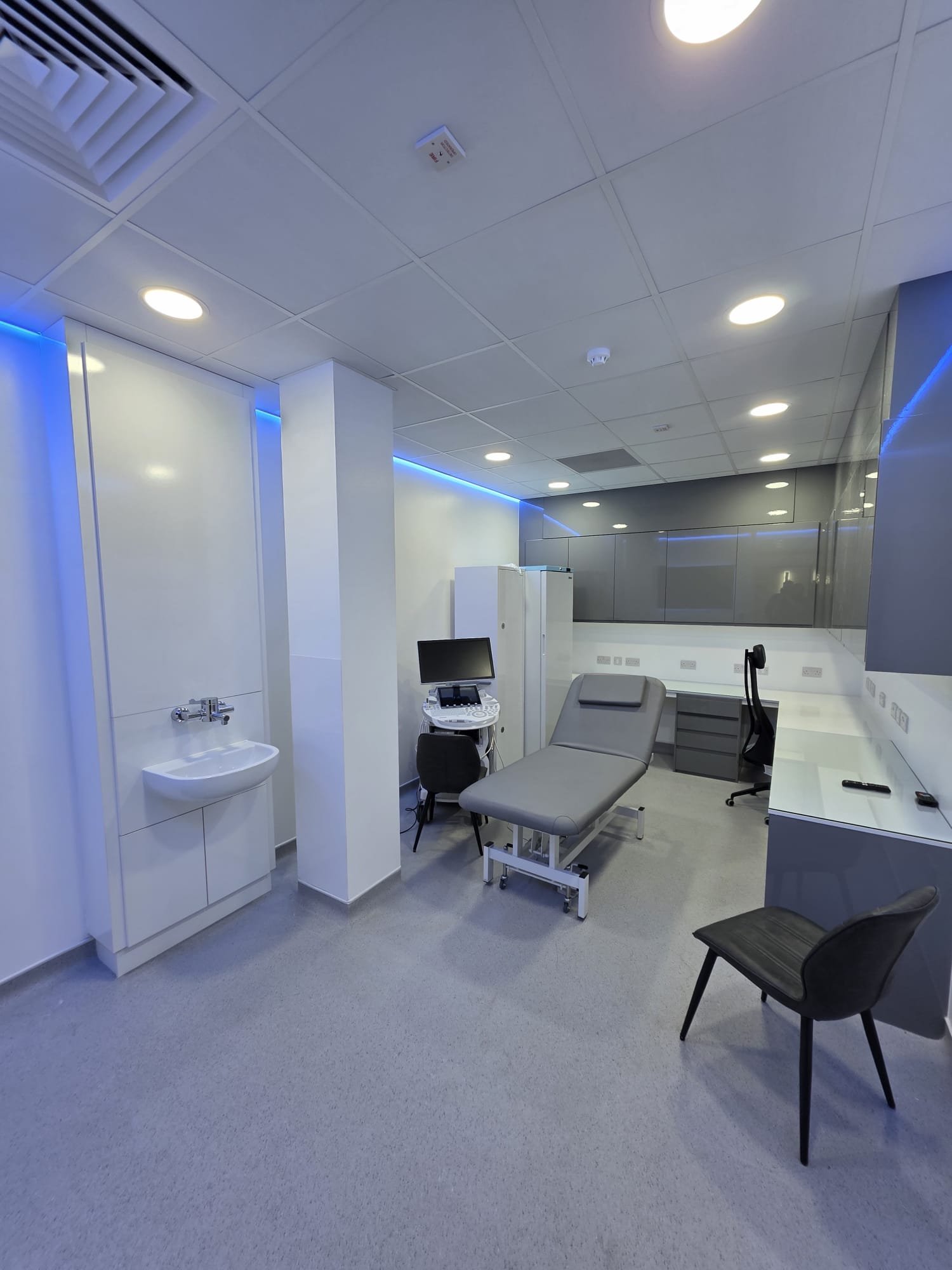 Medical examination room with an examination table, chairs, medical equipment, and monitors, illuminated with ceiling lights and blue accent lighting.