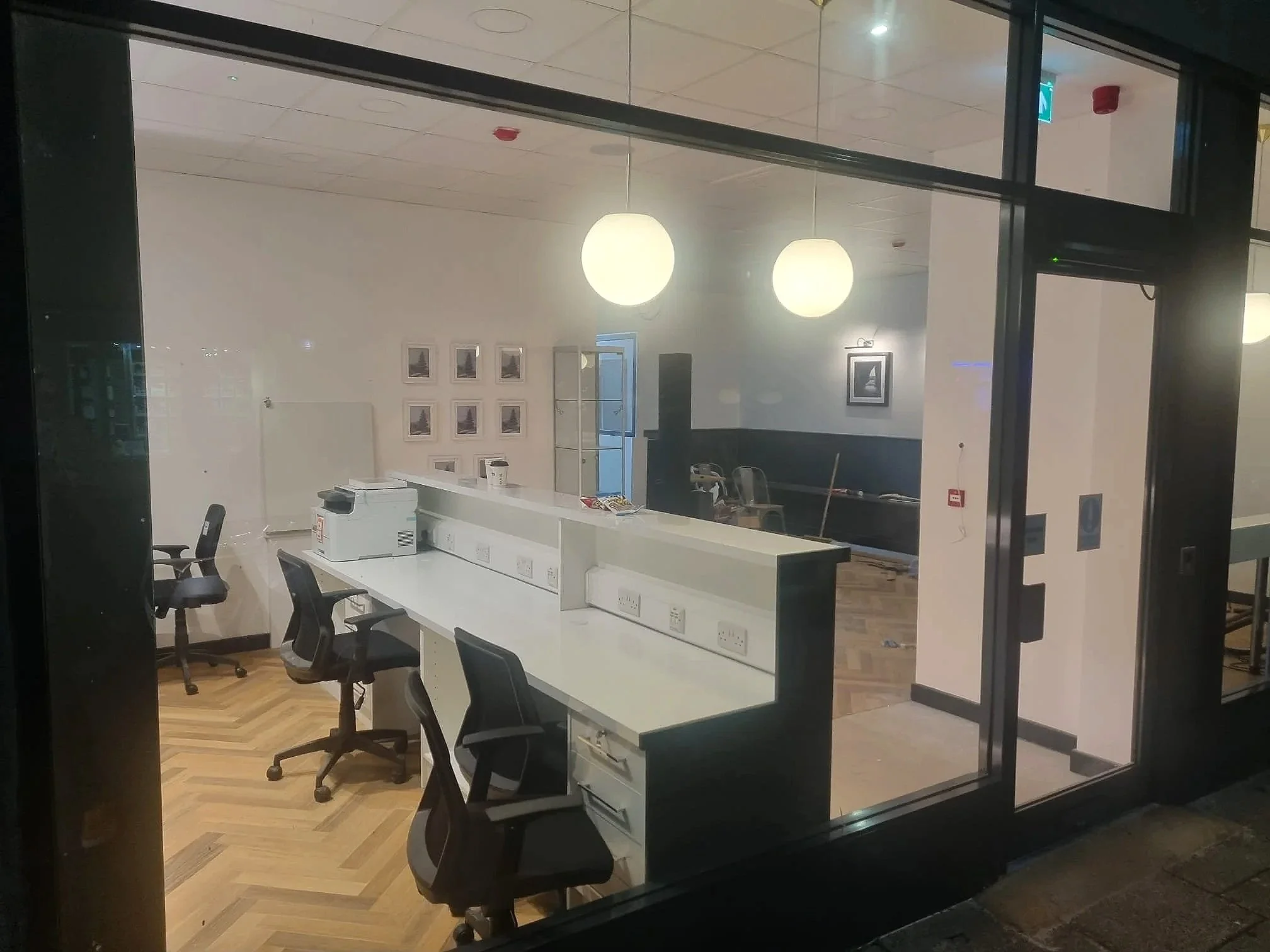 An office or conference room seen through a glass wall and door. The room has a long white table with several black chairs around it, some office equipment such as a printer on the table, framed pictures on the wall, two large round ceiling lights, a