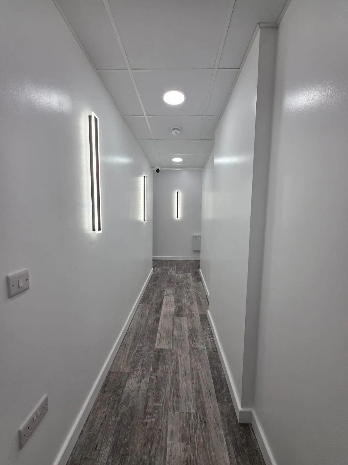 A narrow, white-walled hallway with gray wood-like flooring, illuminated by ceiling lights and wall-mounted LED fixtures.