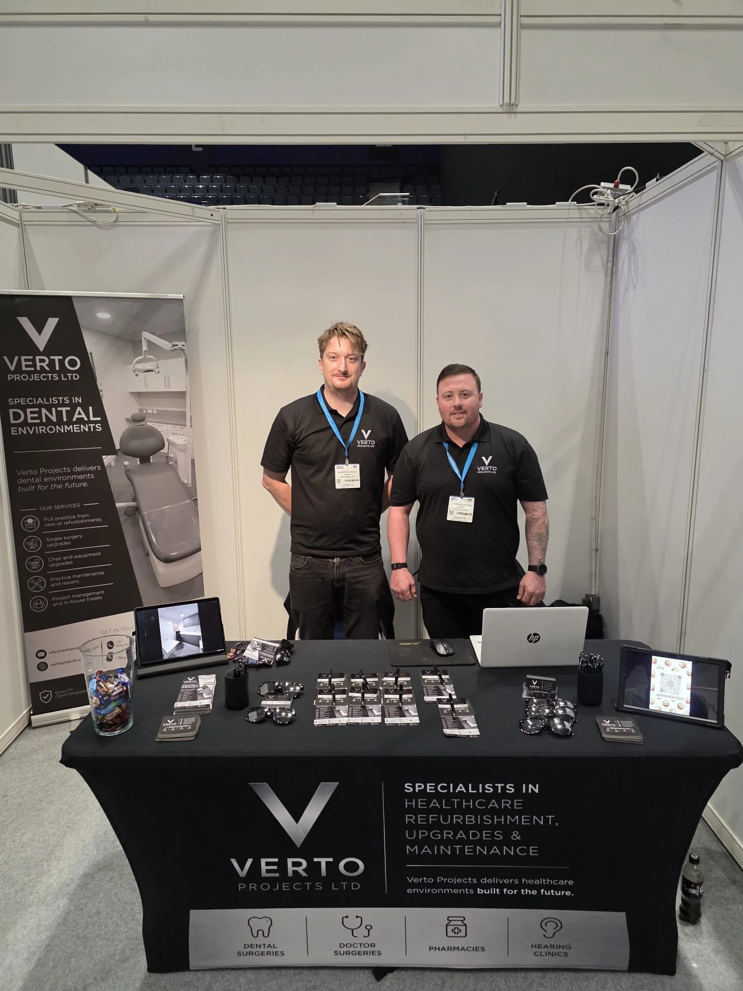 Two men standing behind a booth at a healthcare trade show. The booth advertises Verto Projects Ltd, a company specializing in healthcare refurbishment, upgrades, and maintenance. The booth has promotional materials, electronic devices, and small items like keychains and business cards on the table.