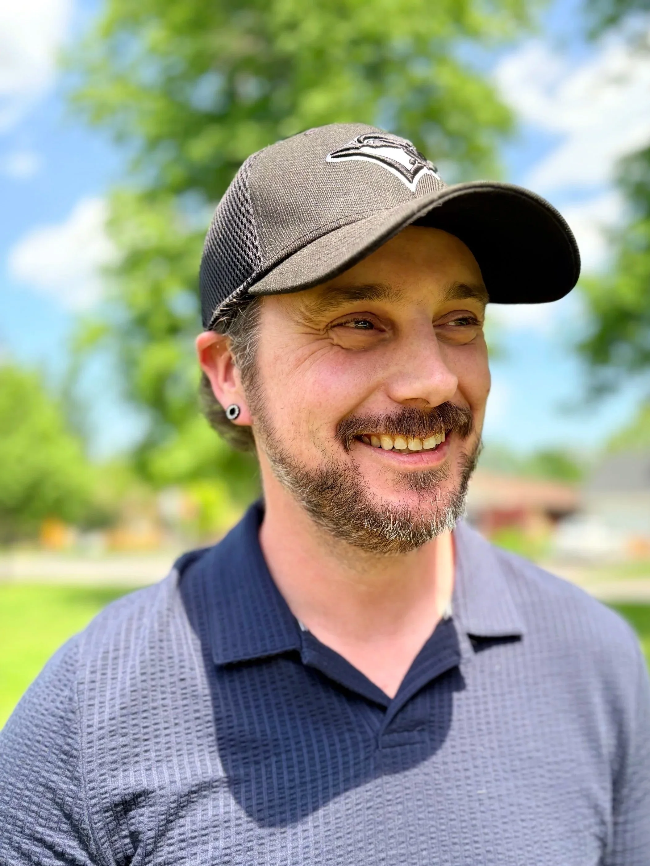 Close-up of David Sutcliffe wearing a gray baseball cap with a sports team logo on it, outdoors with green trees and blue sky in the background.
