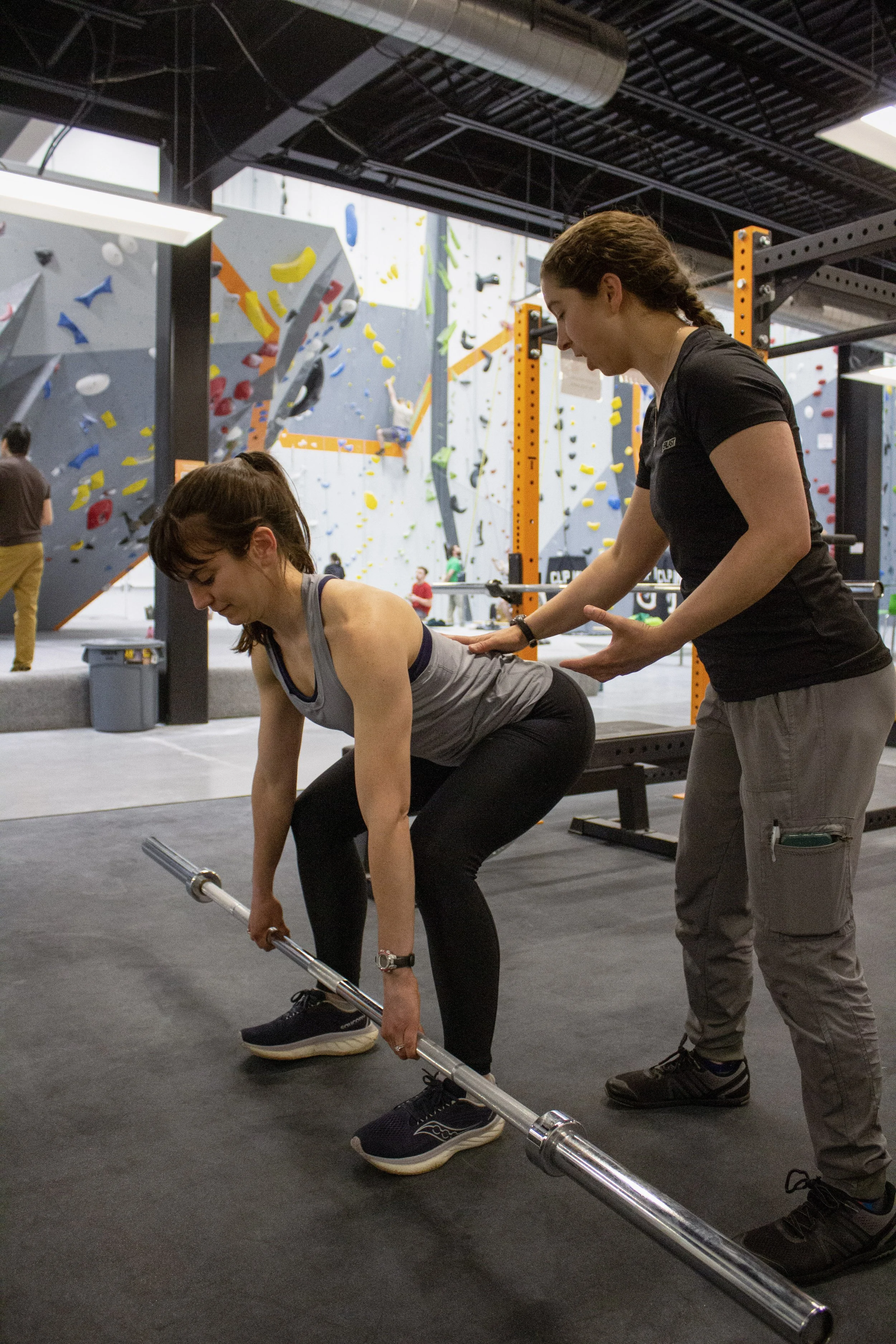 A physical therapist coaches a patient through a barbell deadlift. Rock climbing walls in the background.