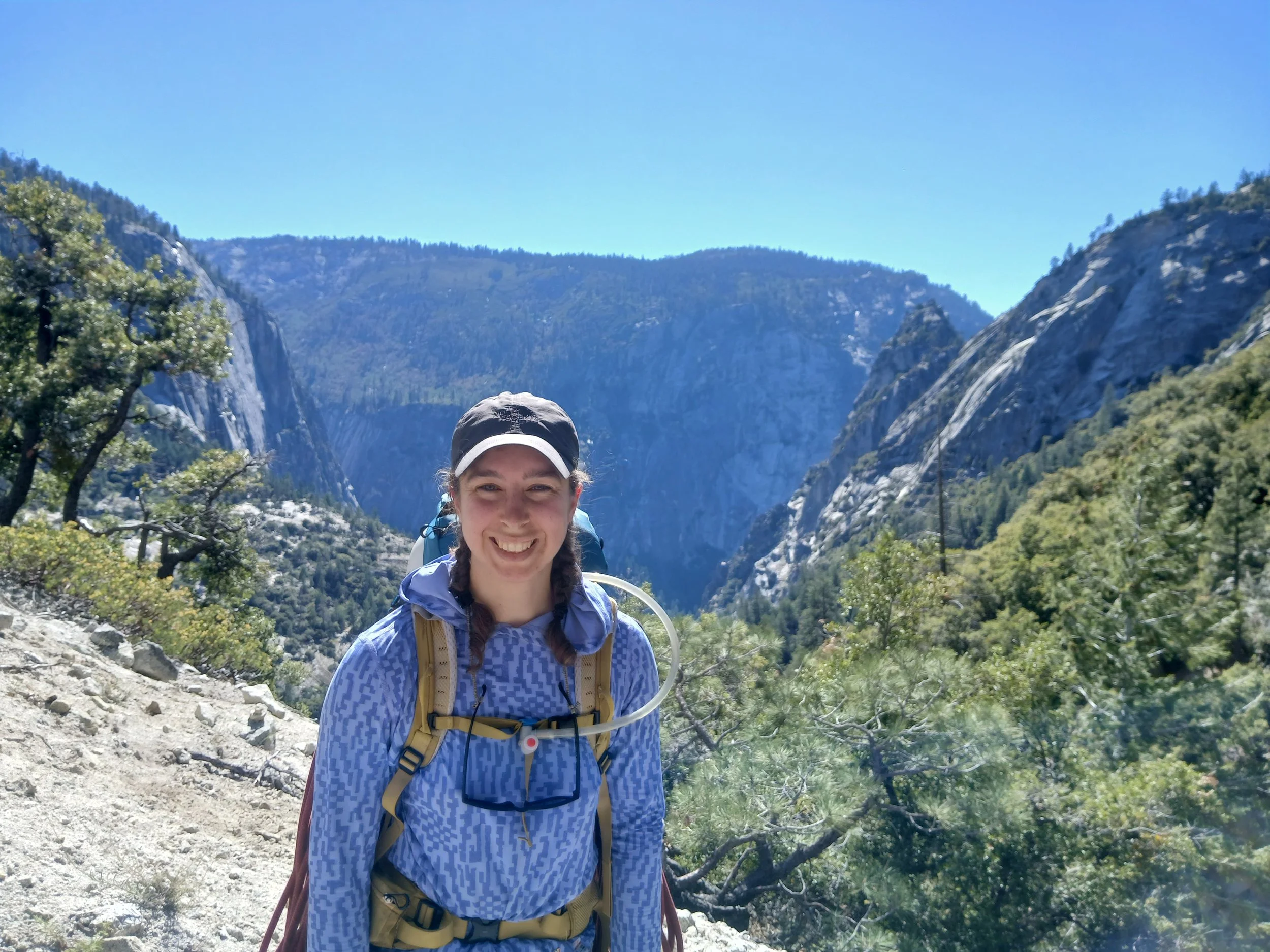 Lori is standing outside on a mountain in the foreground of the picture, in the background there are mountains and blue skies