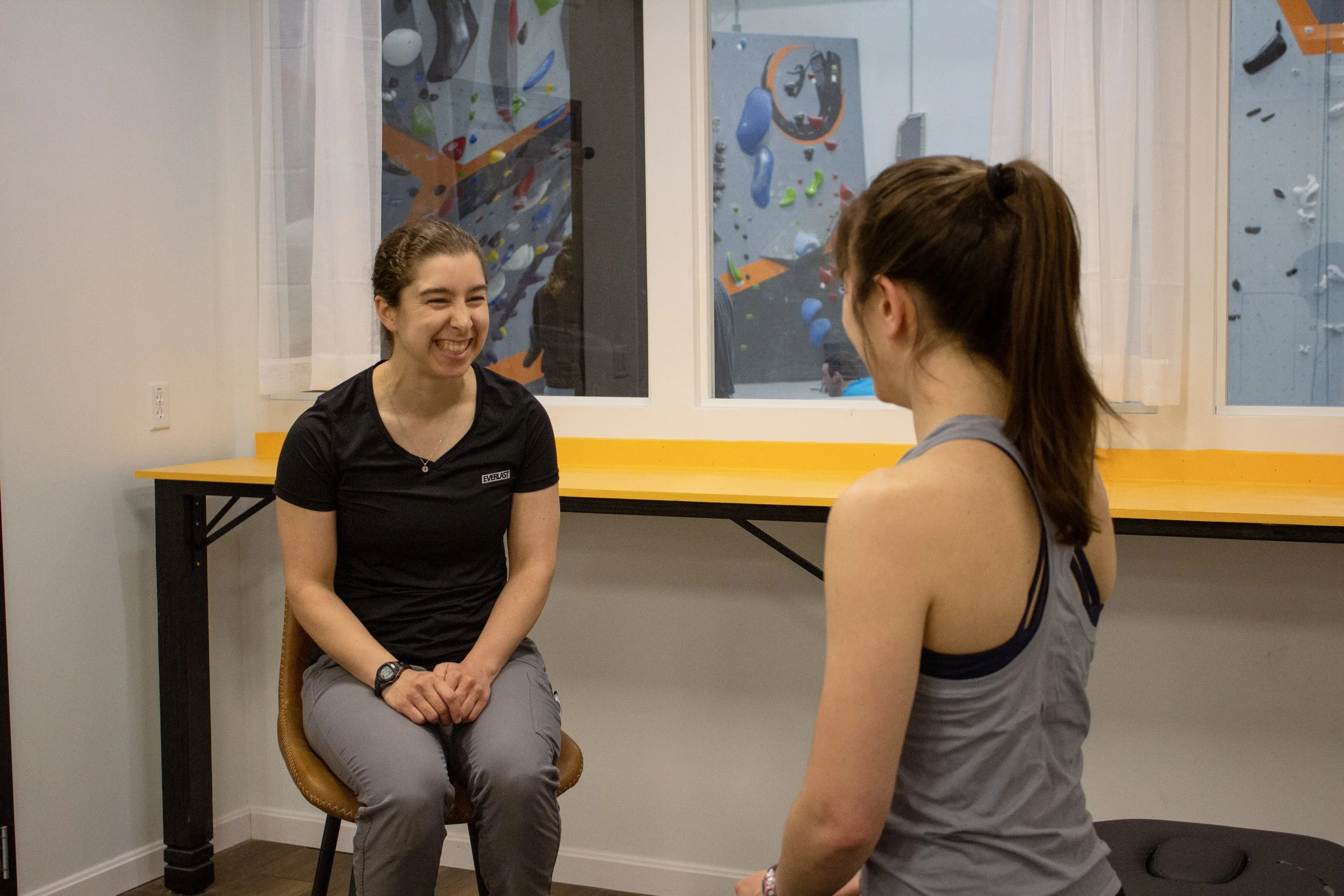 Physical therapist, Lori, sits on a chair speaking with a patient. Lori is laughing in the picture and there are rock climbing walls in the background.