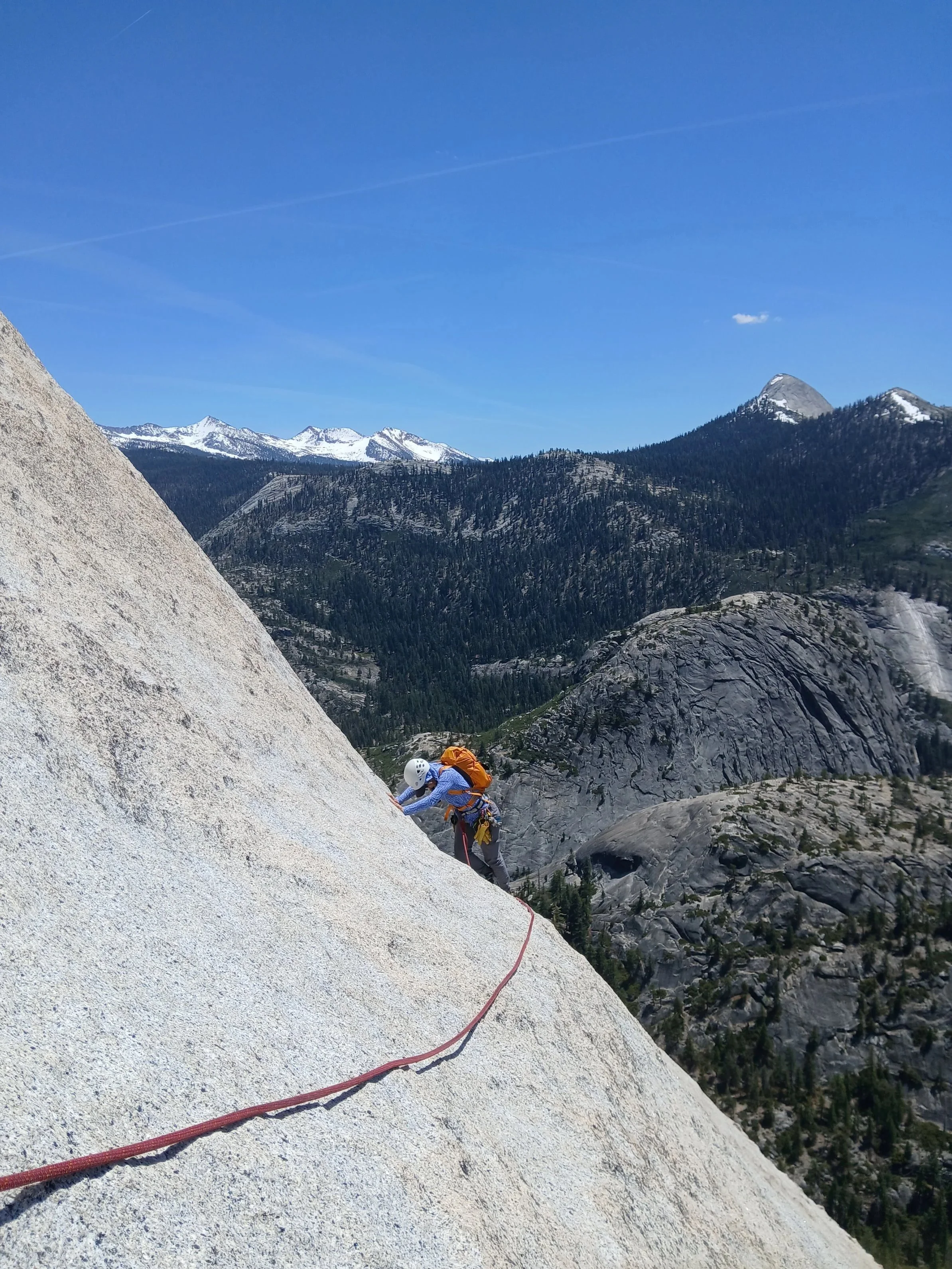 A person wearing a helmet, blue shirt, and orange backpack is rock climbing on a steep granite face in a mountainous area with forested slopes and snow-capped peaks under a clear blue sky.