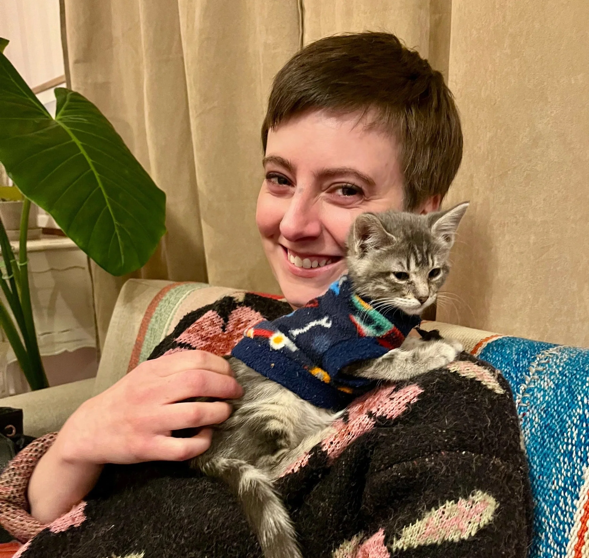 A woman with short brown hair smiling, holding a gray tabby cat wearing a colorful sweater, in a warm-colored living room with a green houseplant and beige curtains.