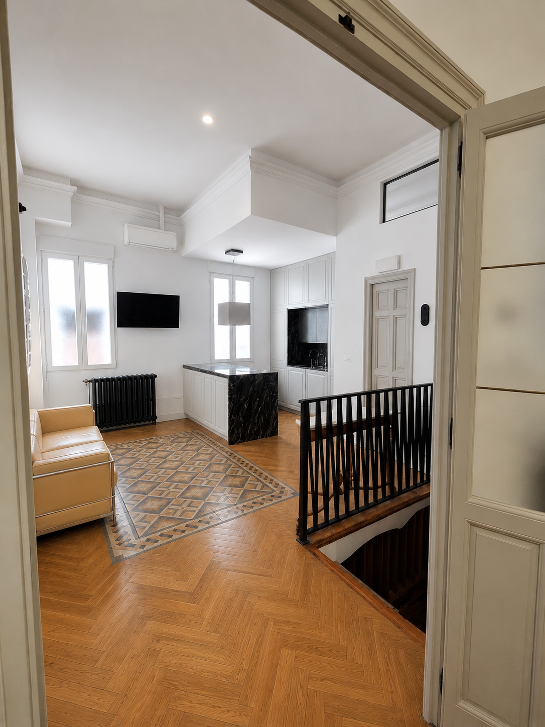 Vista de una sala moderna con suelo de madera, sillón beige, alfombra decorativa, televisión, ventanas y una cocina con isla de mármol negro, barandilla negra y escaleras.