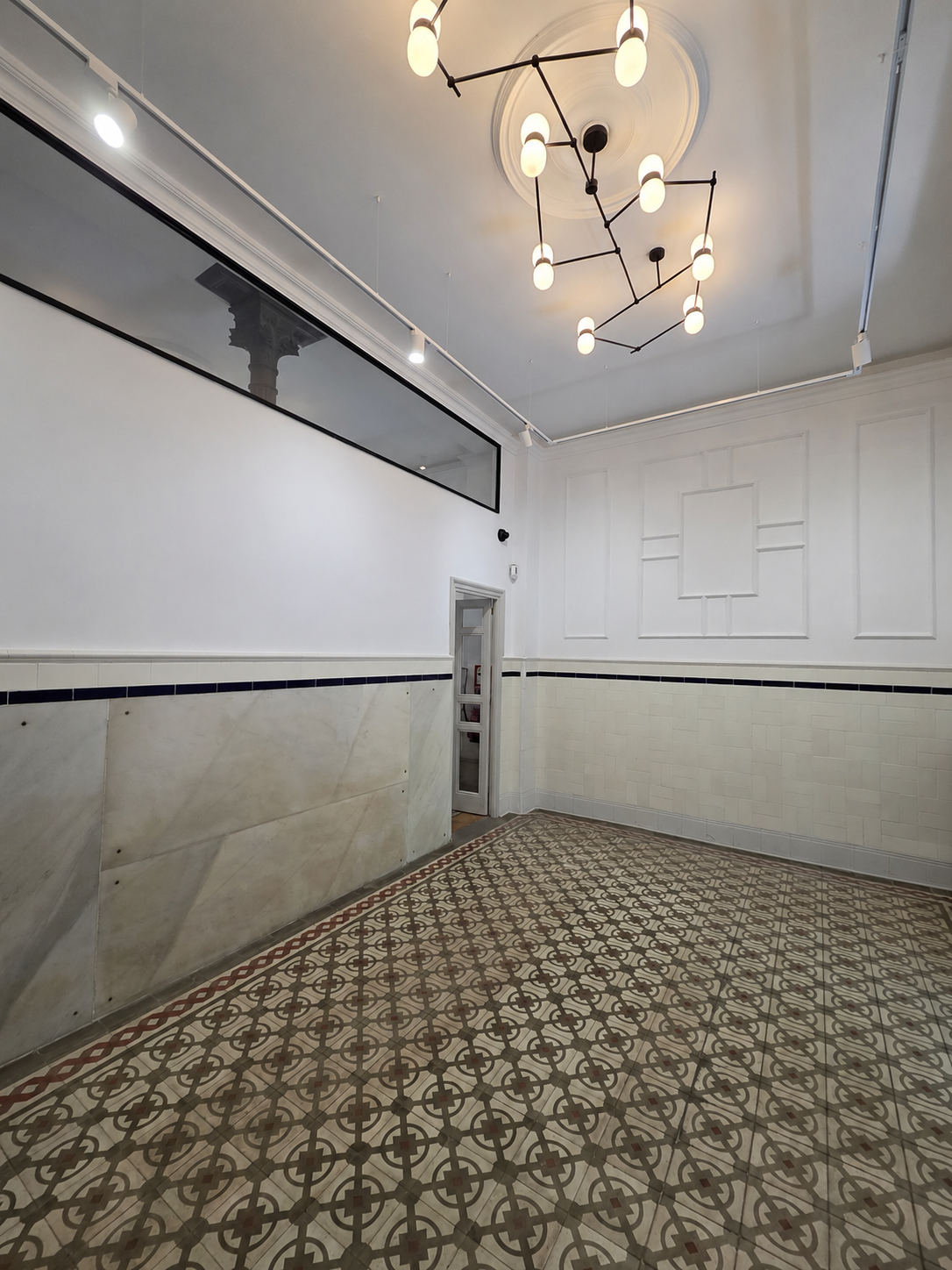 Interior de una sala con paredes blancas, un suelo con baldosas decorativas, un techo con una lámpara moderna y una ventana larga y estrecha.