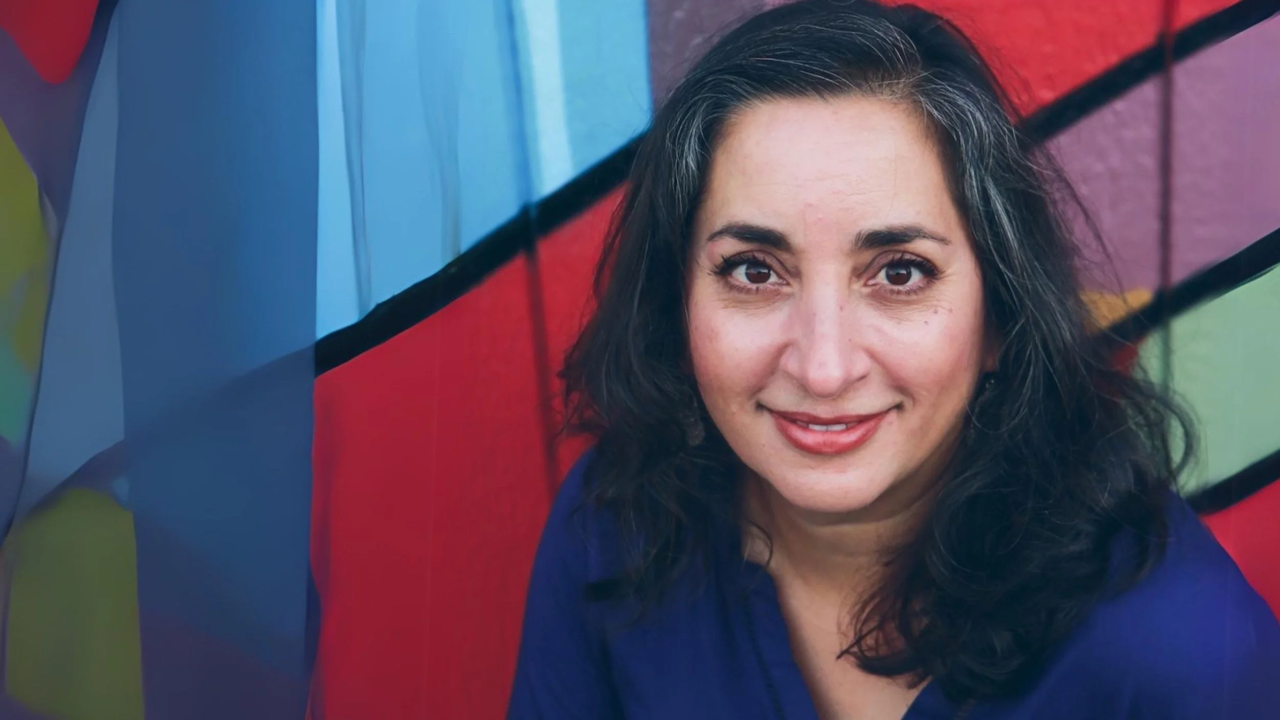 A woman with dark, wavy hair and brown eyes smiling, standing in front of a colorful painted wall.