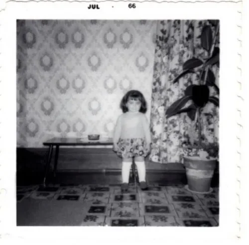 A young girl standing in a living room with vintage wallpaper, a potted plant, and a small table.