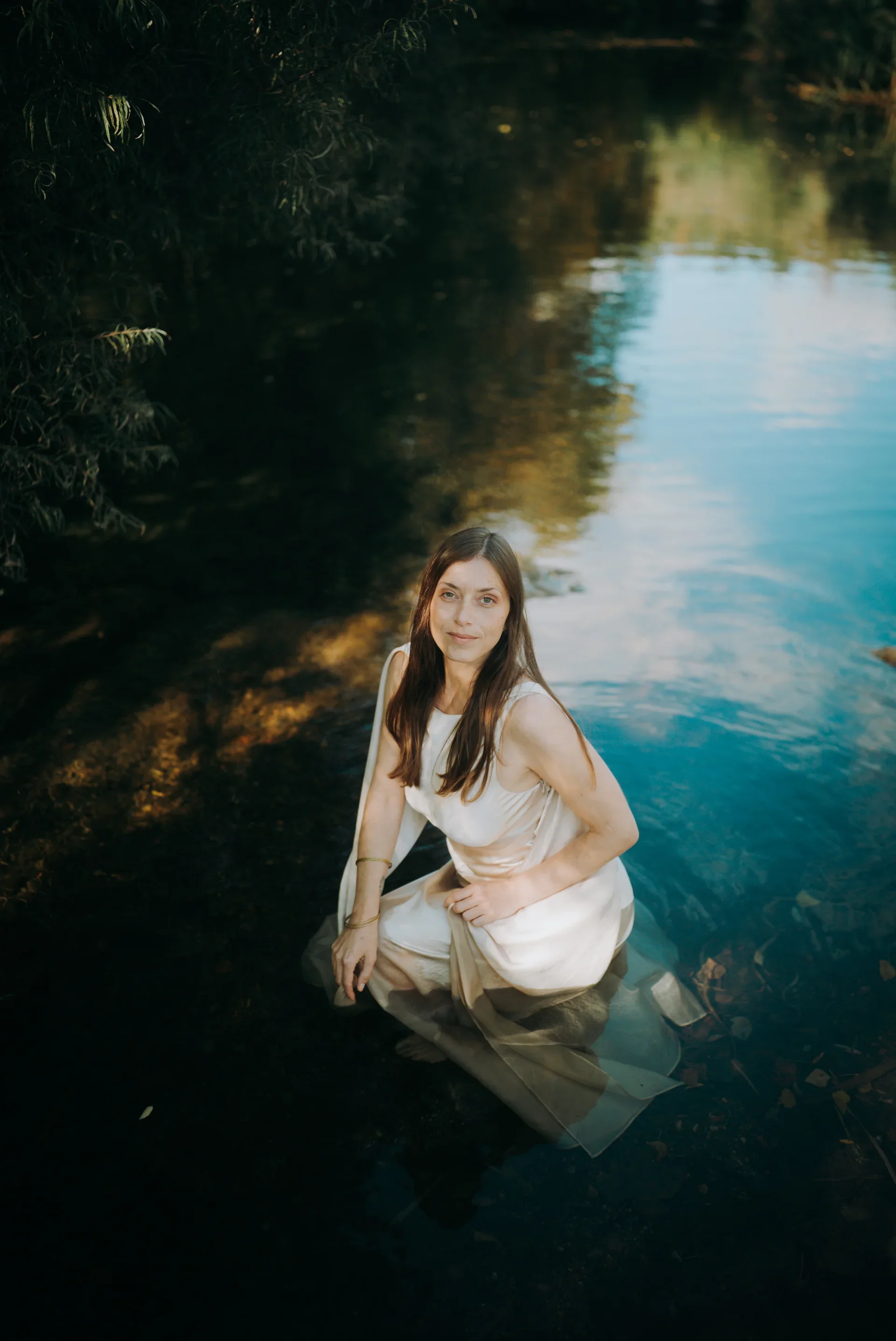 Une femme en robe blanche est assise dans l'eau d'une rivière ou d'un lac avec des arbres et la nature en arrière-plan, l'eau est claire et reflète le paysage.