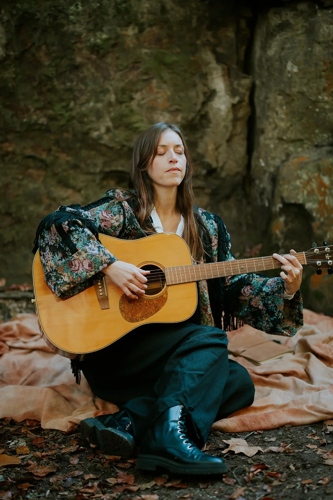 Une femme assise sur le sol, jouant de la guitare acoustique, dans un cadre naturel avec des rochers et des feuilles mortes.