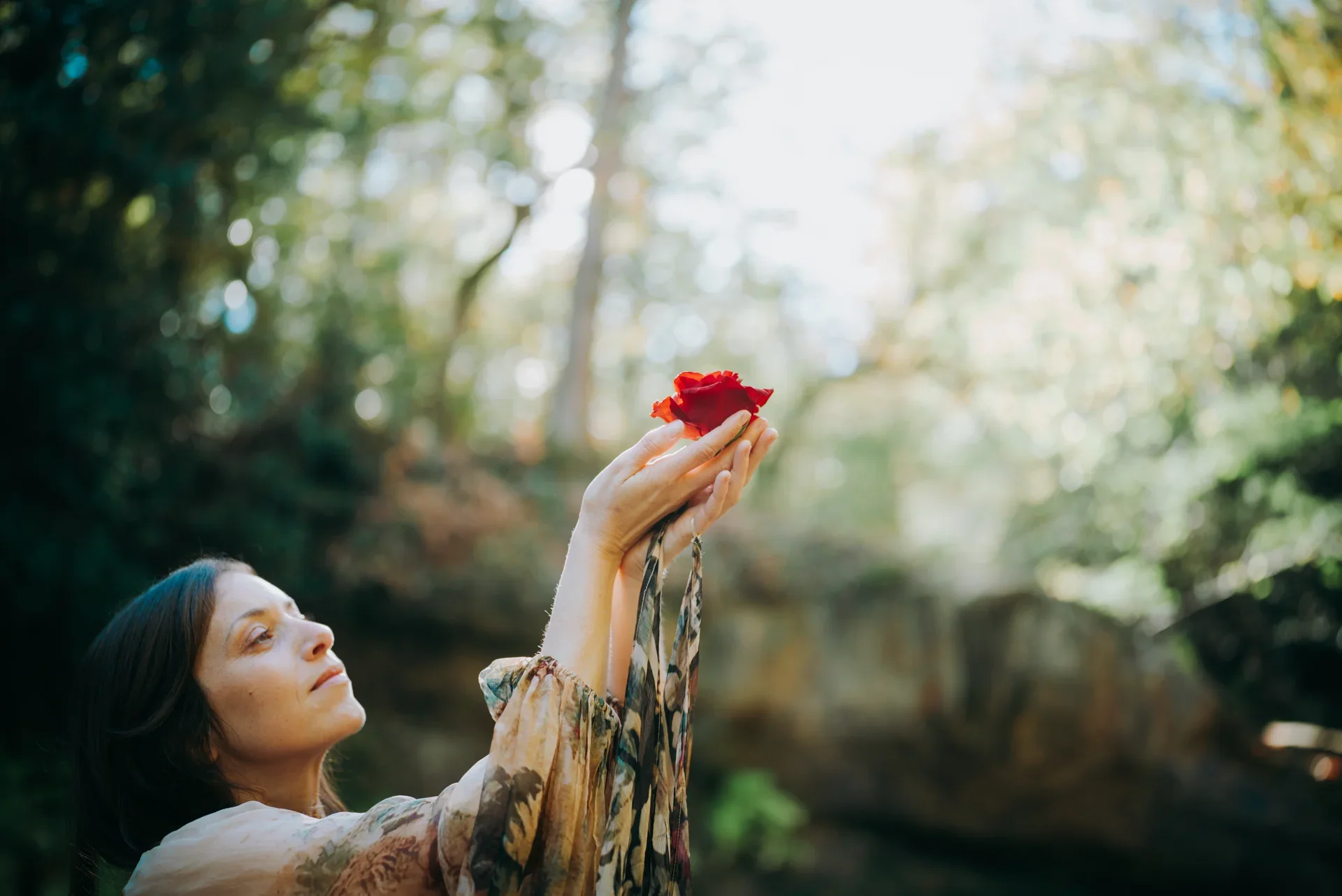 Une femme avec des cheveux bruns tient une rose rouge en main dans un environnement naturel avec des arbres et de la lumière douce.