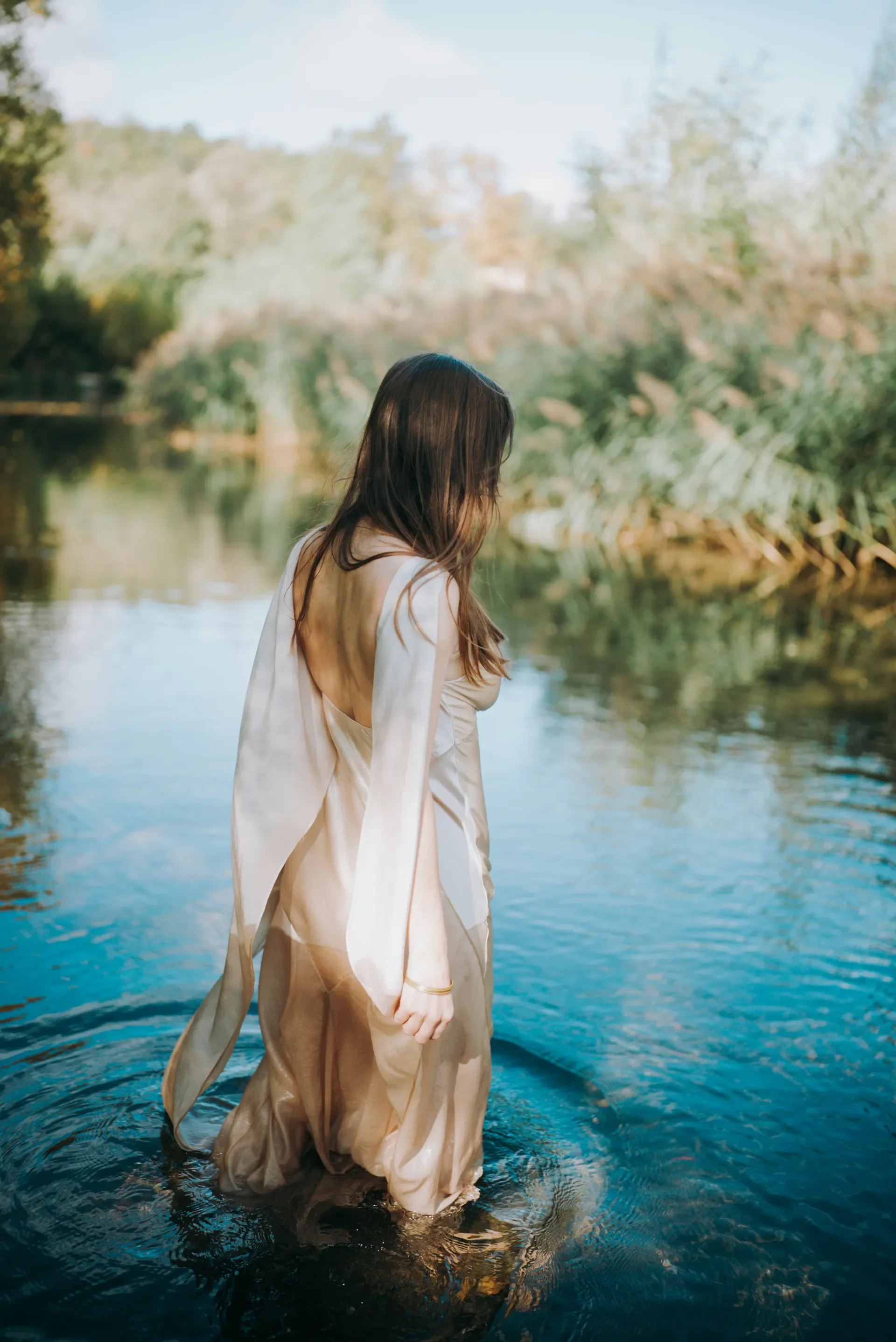 Une femme aux cheveux longs et bruns en robe légère, debout dans une rivière, entourée de verdure et de végétation, avec un ciel clair en arrière-plan.