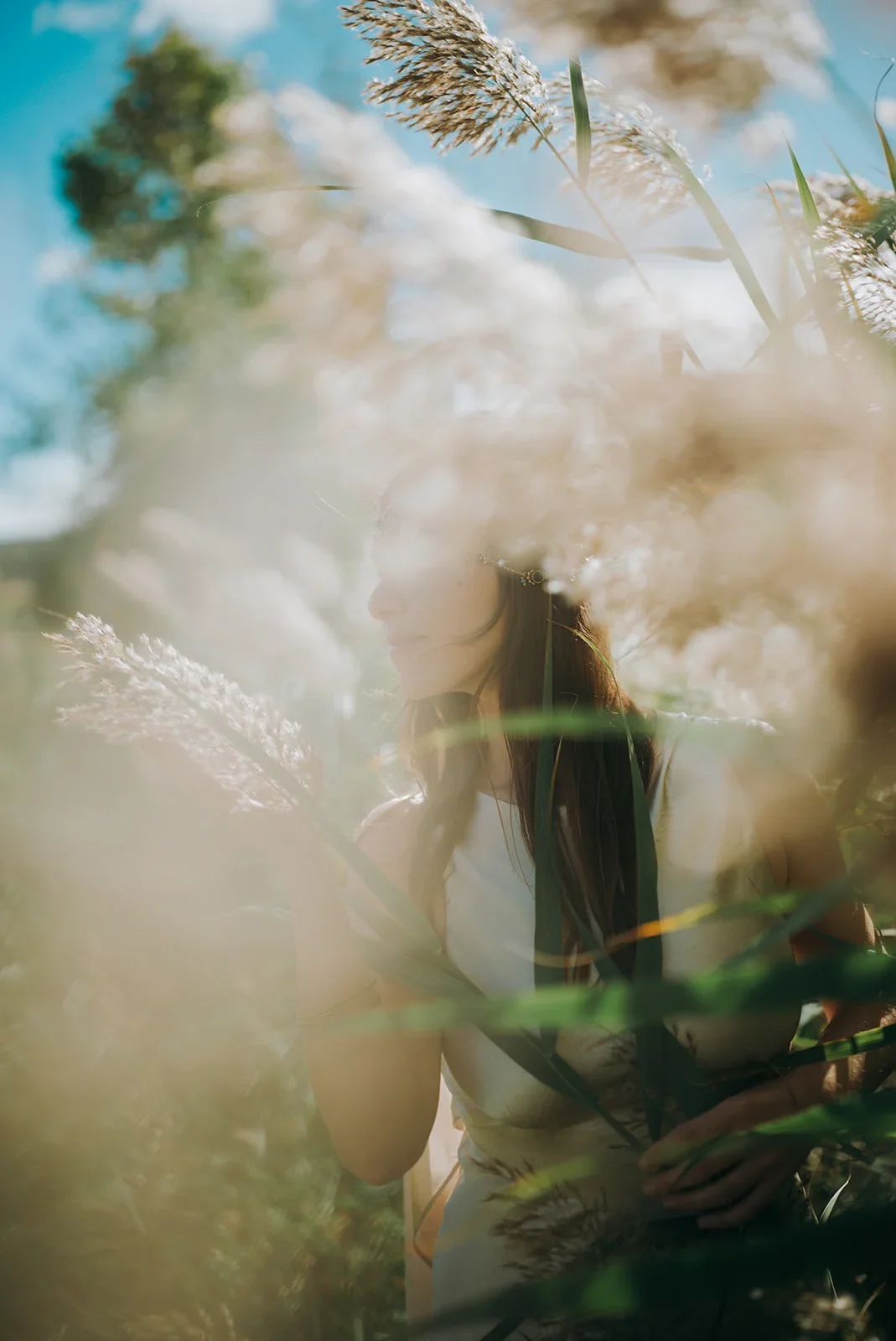 Une femme debout dans un champ herbeux, entourée de plantes hautes et de graminées, avec le ciel bleu en arrière-plan, vue partielle avec mise au point douce et lumière naturelle.