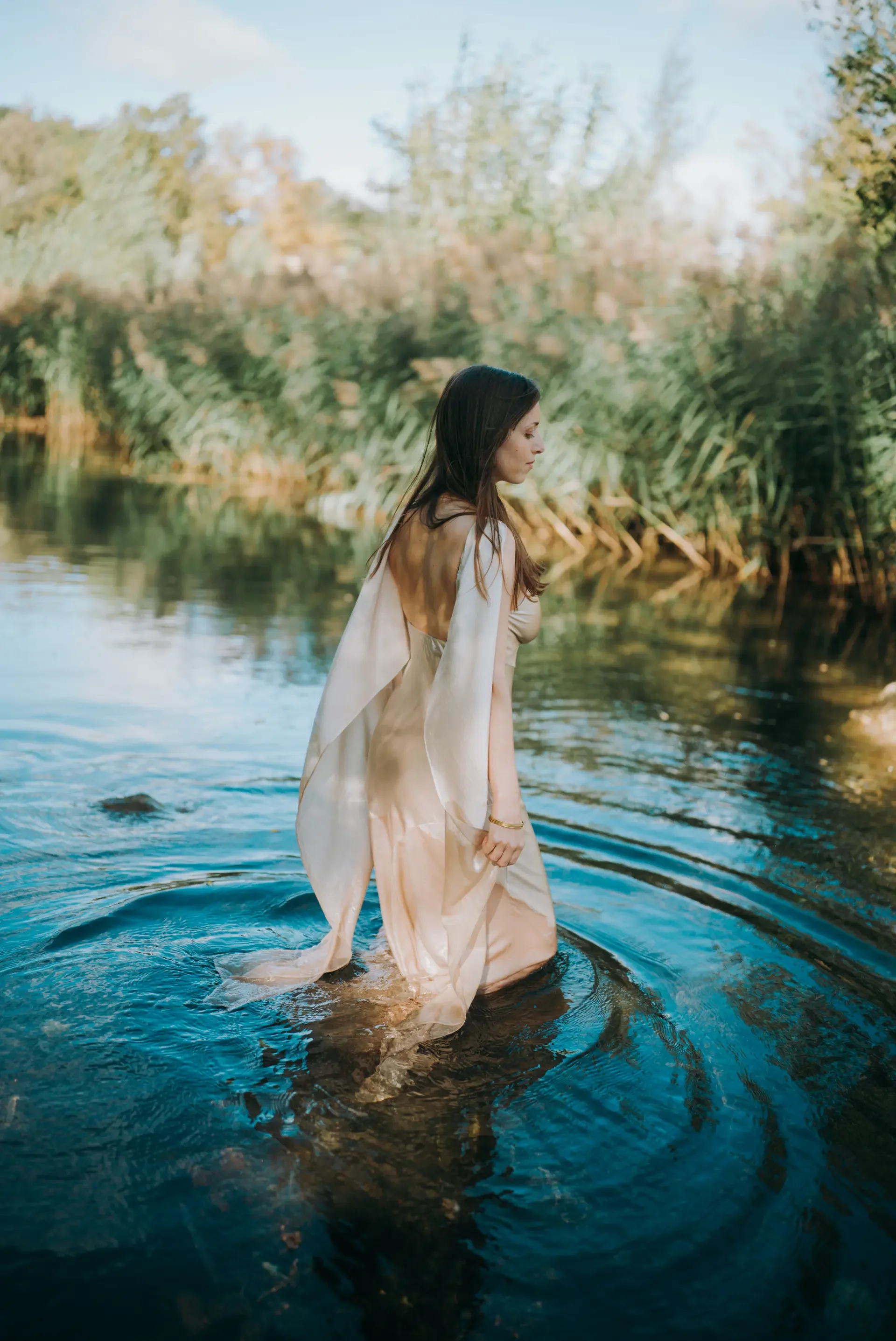 Une femme en robe légère marche dans une rivière, entourée de végétation luxuriante et de nature sauvage.