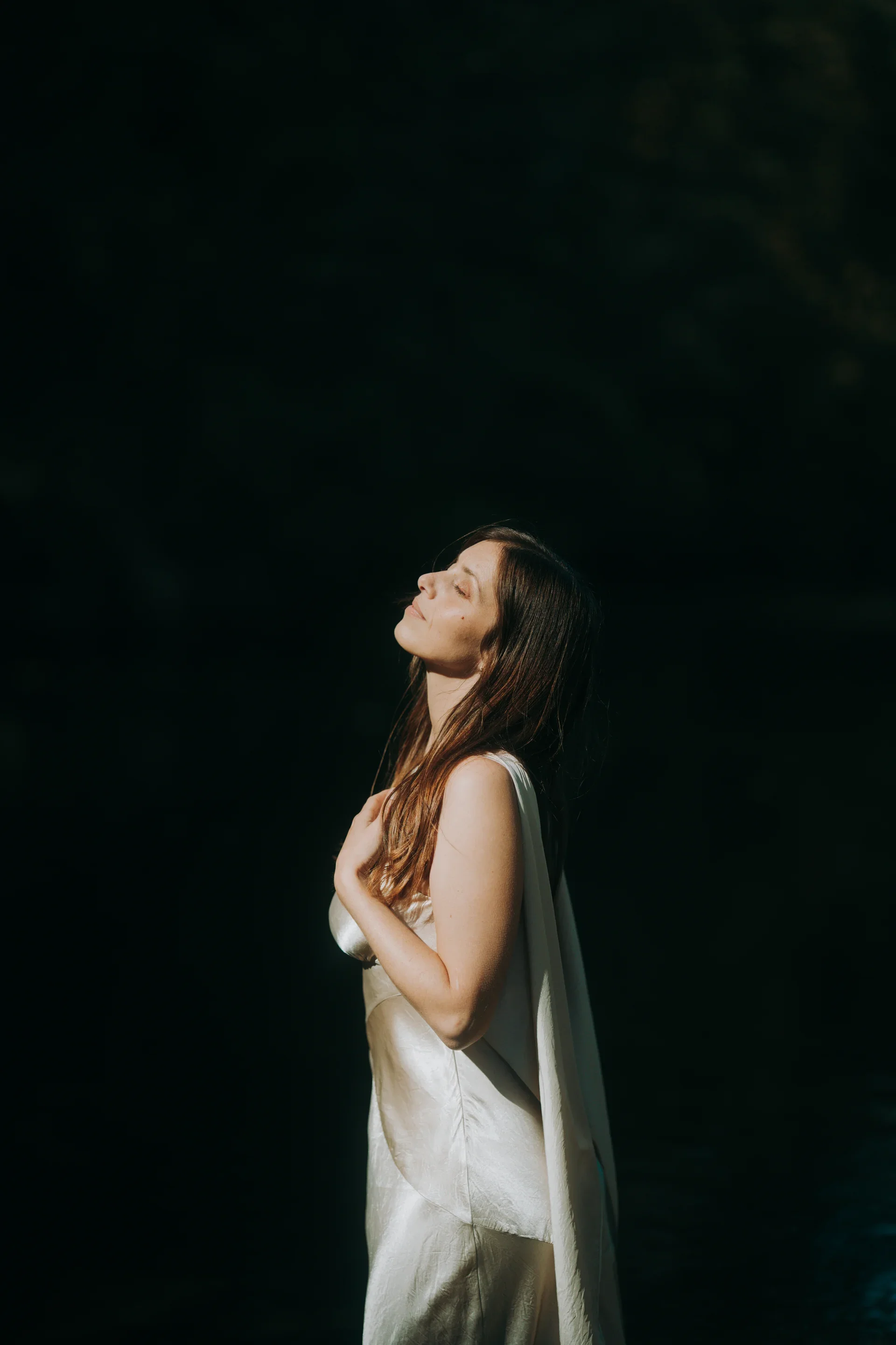 Une femme avec des cheveux longs, vêtue d'une robe en satin crème, se tient les yeux clos, dans une lumière douce contre un fond noir.