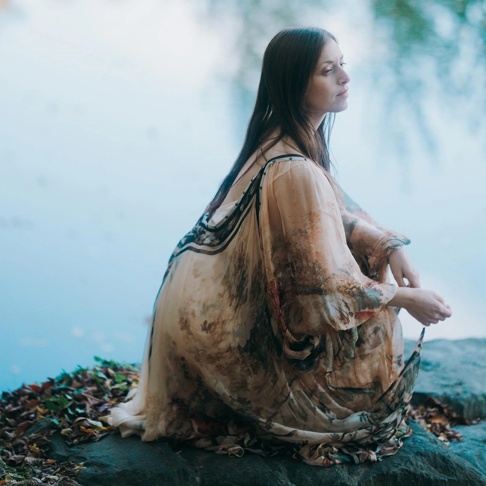 Une femme aux cheveux longs et foncés, assise près d'un plan d'eau, portant une robe légère de couleur beige avec des motifs floraux, regarde pensivement vers l'eau.