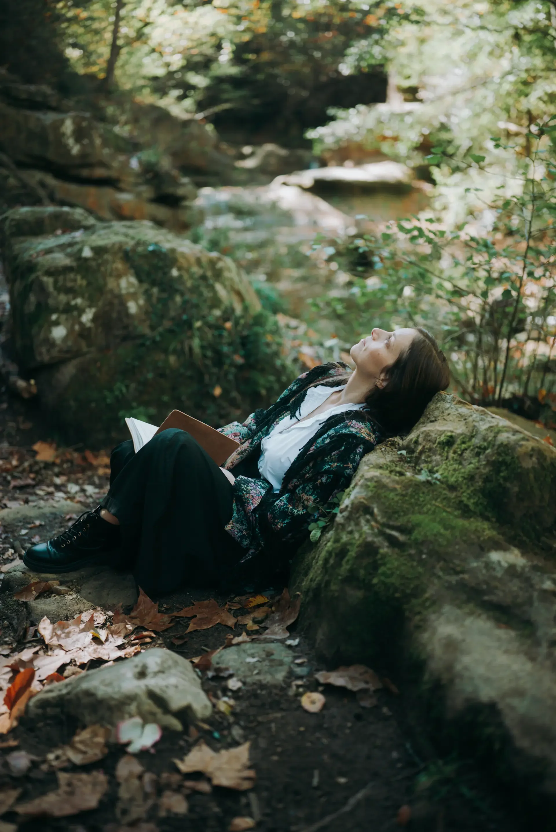 Une femme assise contre une roche dans la forêt, les yeux fermés, en train de se détendre avec un carnet sur ses genoux, entourée d'arbres et de feuilles tombées.