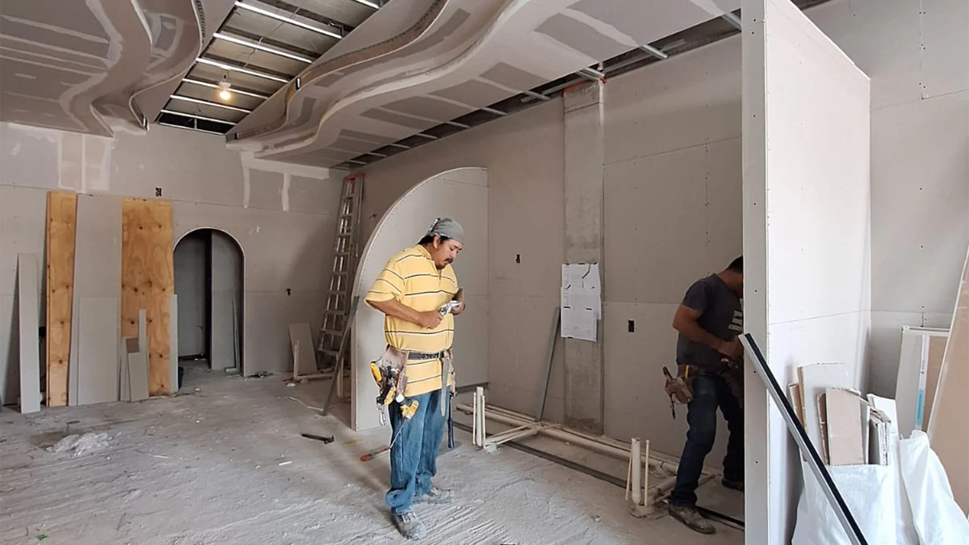 Pareja de trabajadores en una construcción, con herramientas en un espacio en remodelación, con techos en proceso de instalación y paredes de drywall.