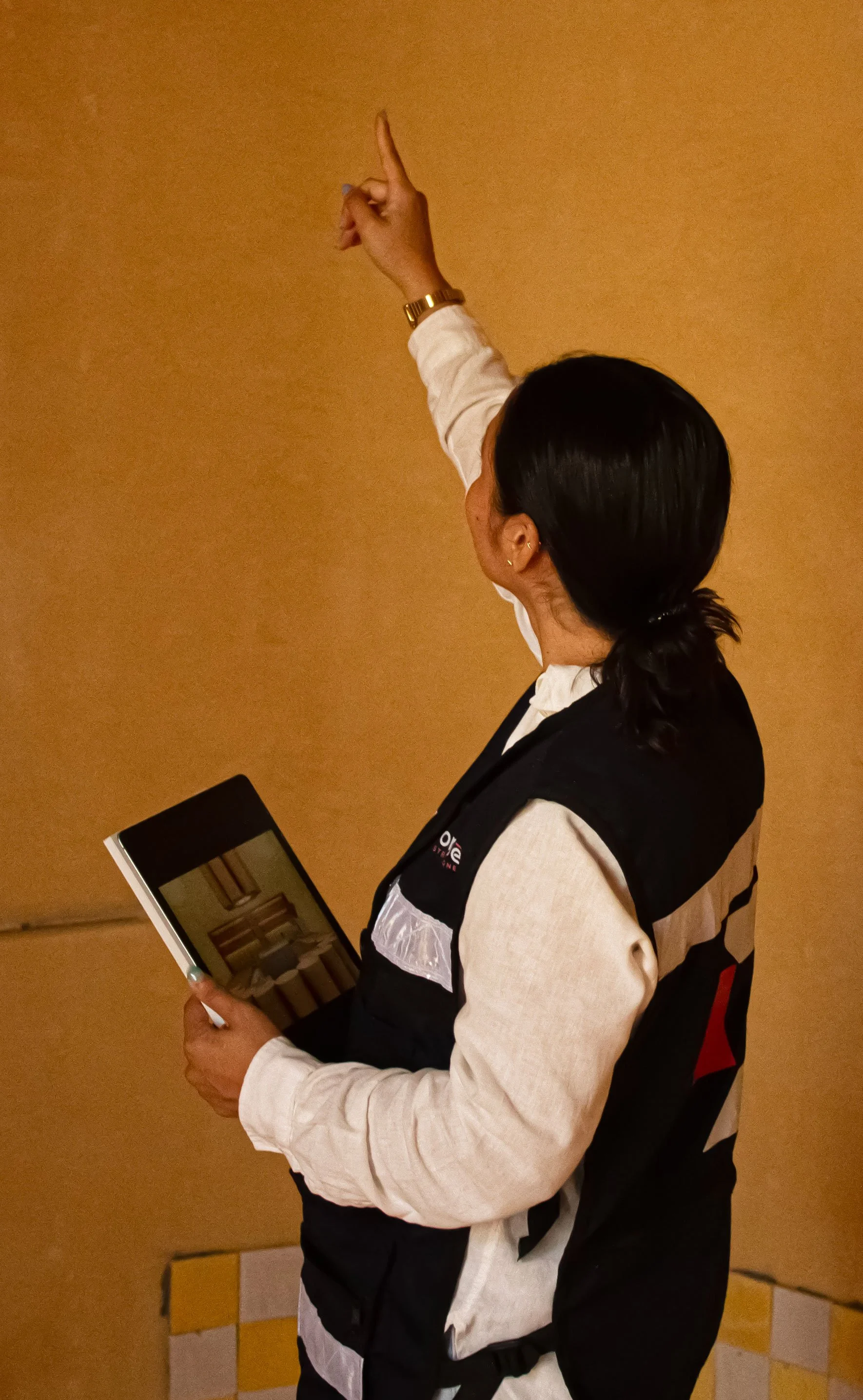 Mujer con ropa de colores claros y oscuro, con cabello oscuro recogido, sosteniendo una tableta y señalando con el dedo hacia arriba, en un fondo de pared color mostaza.