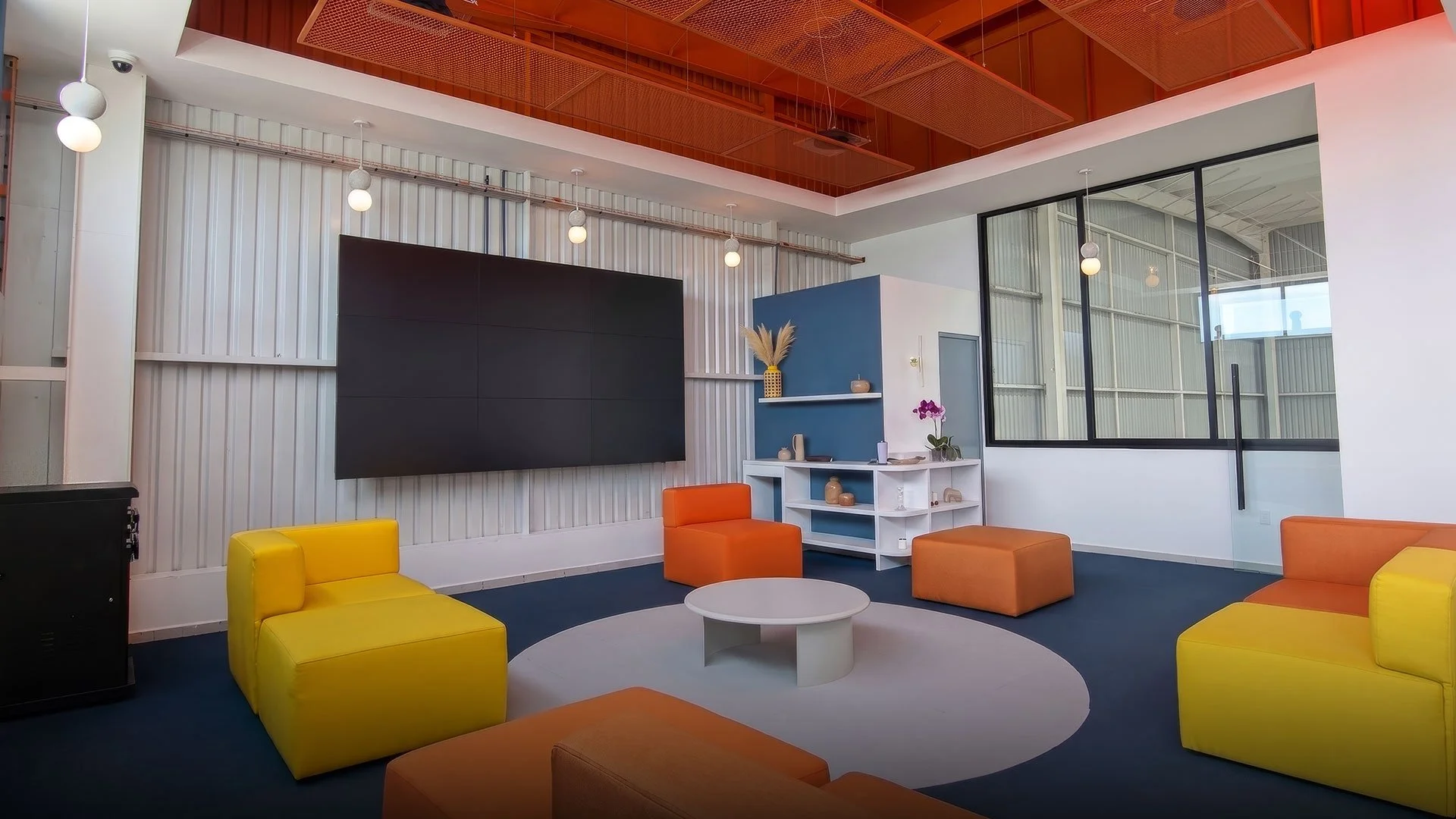 Sala moderna con sillones en colores naranja y amarillo, una mesa blanca redonda, estantería con decoraciones, paredes blancas con un tablero negro y gran ventana con vista a un edificio.
