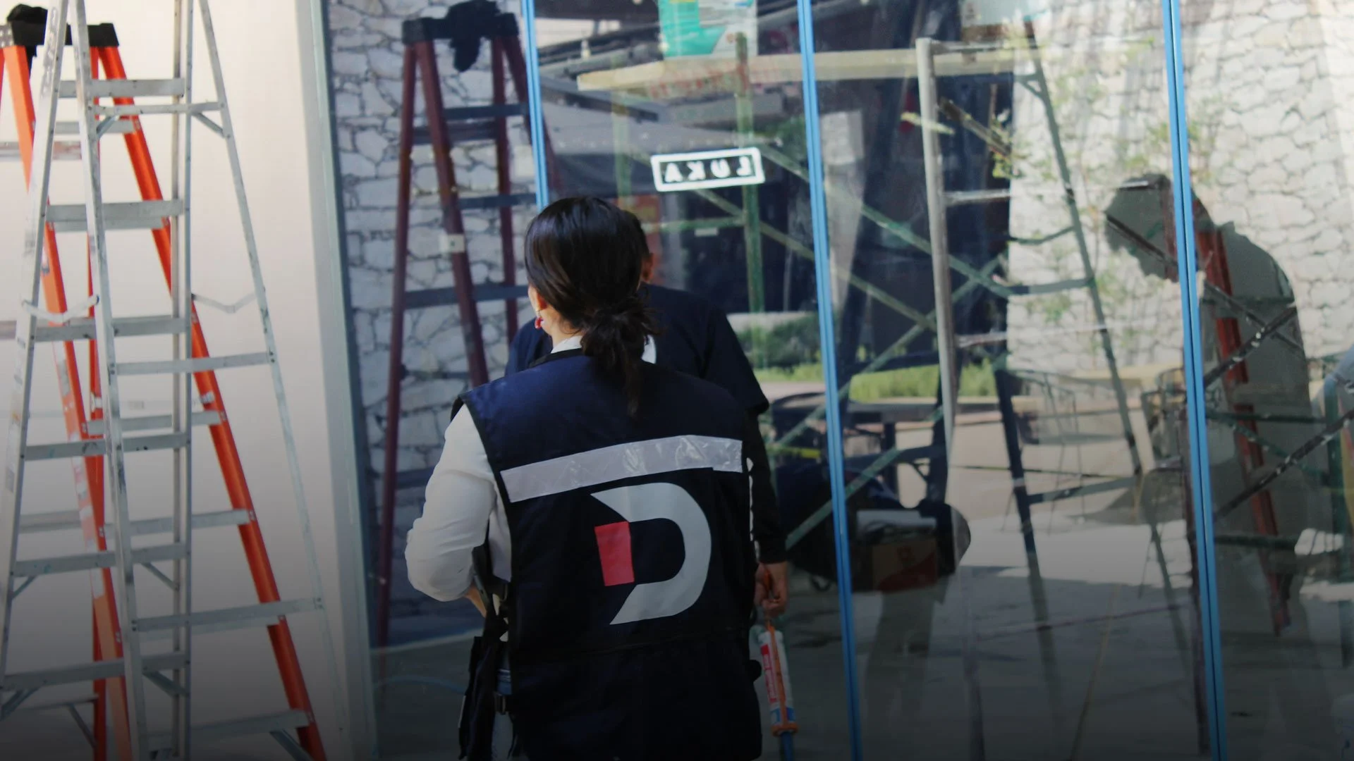 Persona con uniforme de trabajo frente a un vidrio con reflejo, junto a escaleras de aluminio y escaleras de madera en un entorno de construcción o reparaciones.