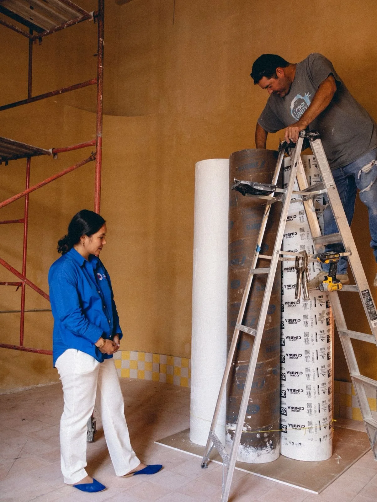 Hombre en escaleras trabajando con un tambor y una mujer observando, en un espacio con paredes beige y estructura de andamio roja.