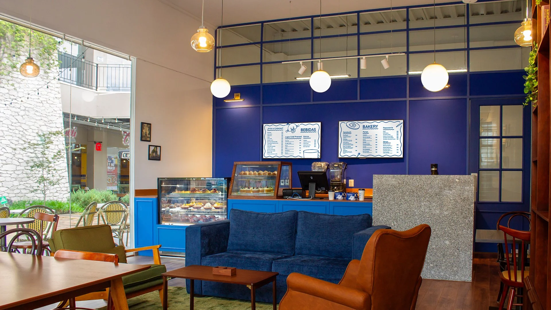 Interior de cafetería con sofá azul, mesas de madera y vitrina con postres, cartel de menú digital en una pared azul, ventanas grandes y plantas decorativas.