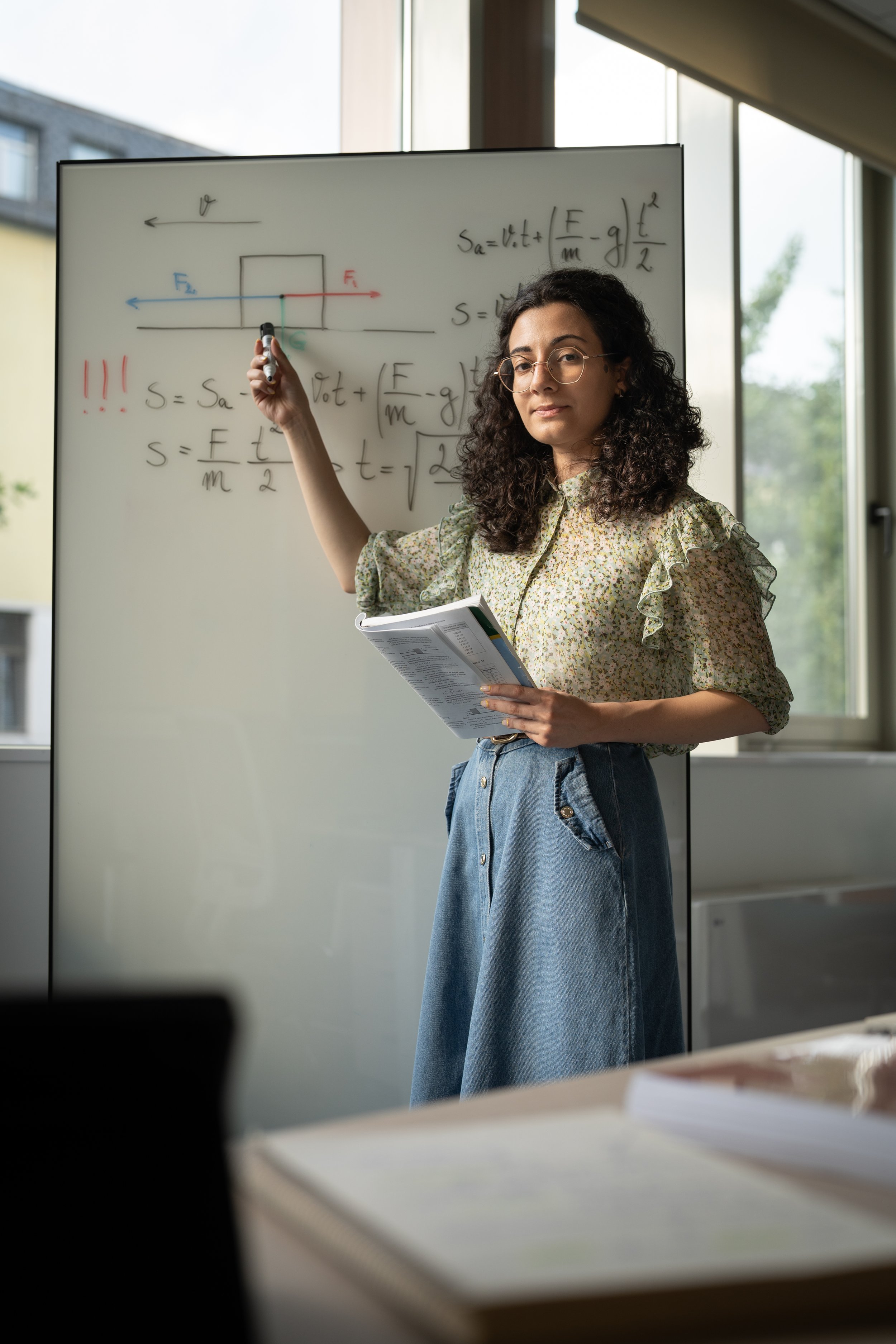 A woman with curly dark hair, glasses, and a floral blouse writing formulas and diagrams about physics or mechanics on a whiteboard in a classroom or office with large windows.
