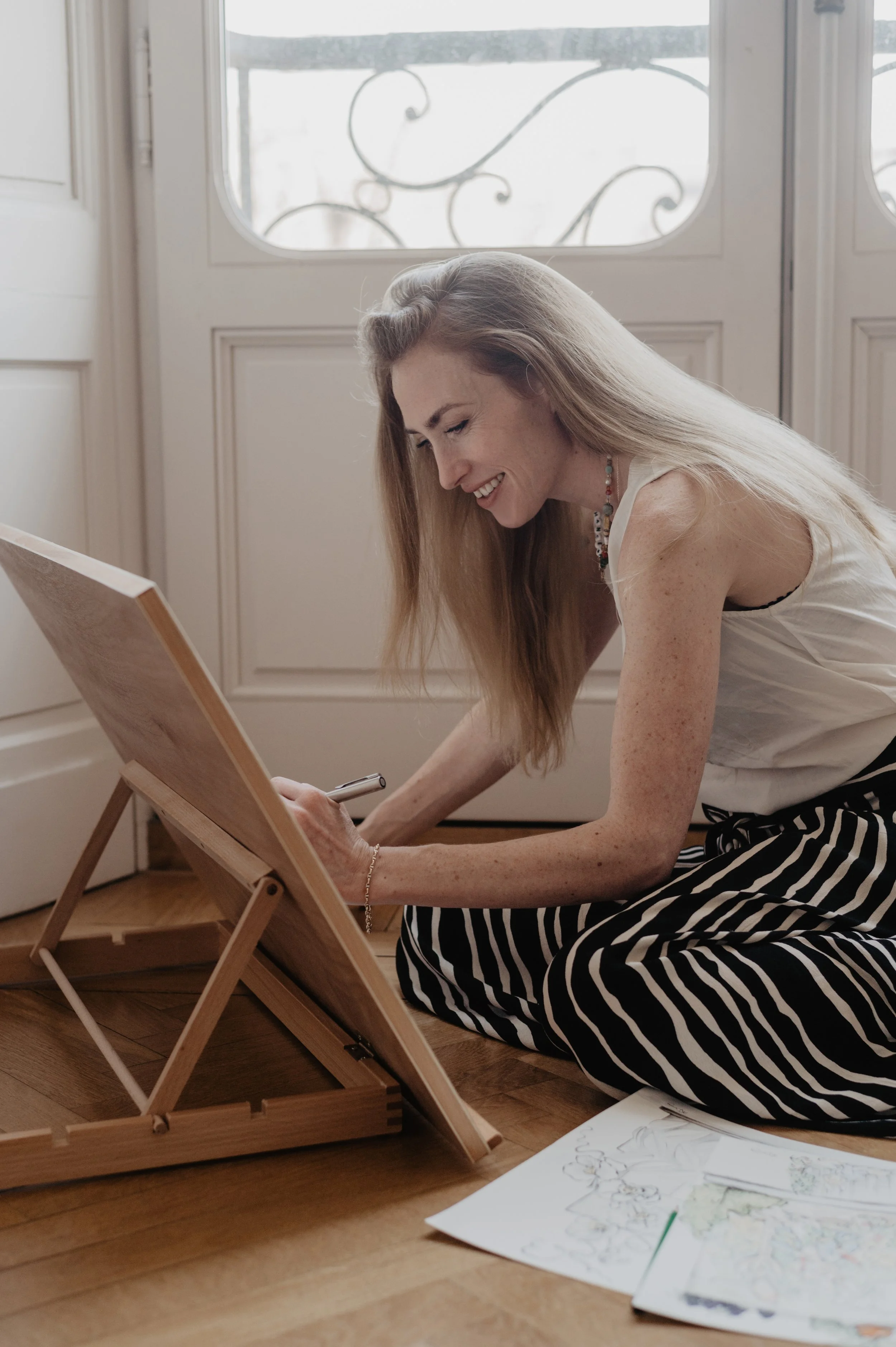 A woman with long blonde hair is smiling and drawing on a large sheet of paper. The paper is propped on a small wooden easel on the floor. She is sitting on the wooden floor with her legs crossed and wearing a striped skirt and a sleeveless top near a white door with decorative ironwork outside.