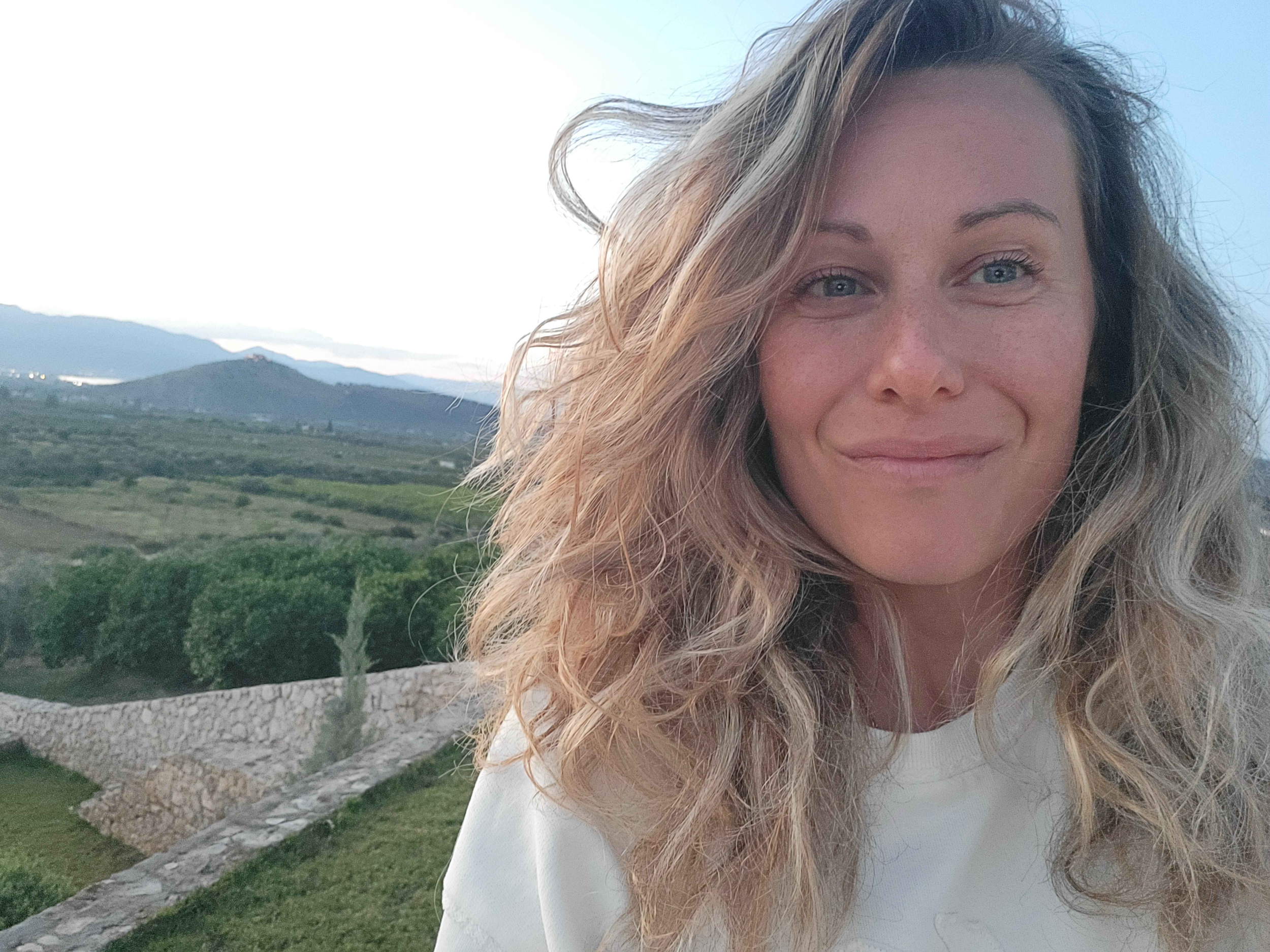 Une femme aux cheveux bouclés et aux yeux bleus sourit devant un paysage rural avec des montagnes au loin, un ciel bleu et des collines verdoyantes.