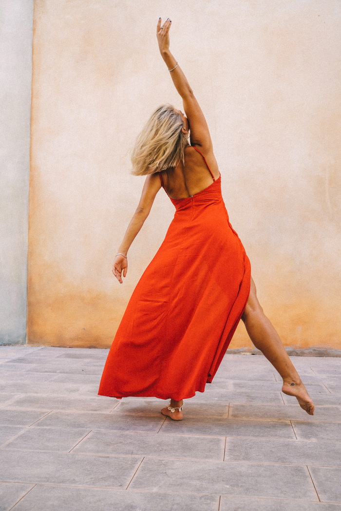 Femme danse en robe longue rouge, pied nu, devant un mur beige, bras levé, cheveux blonds mi-longs, mouvement fluide.
