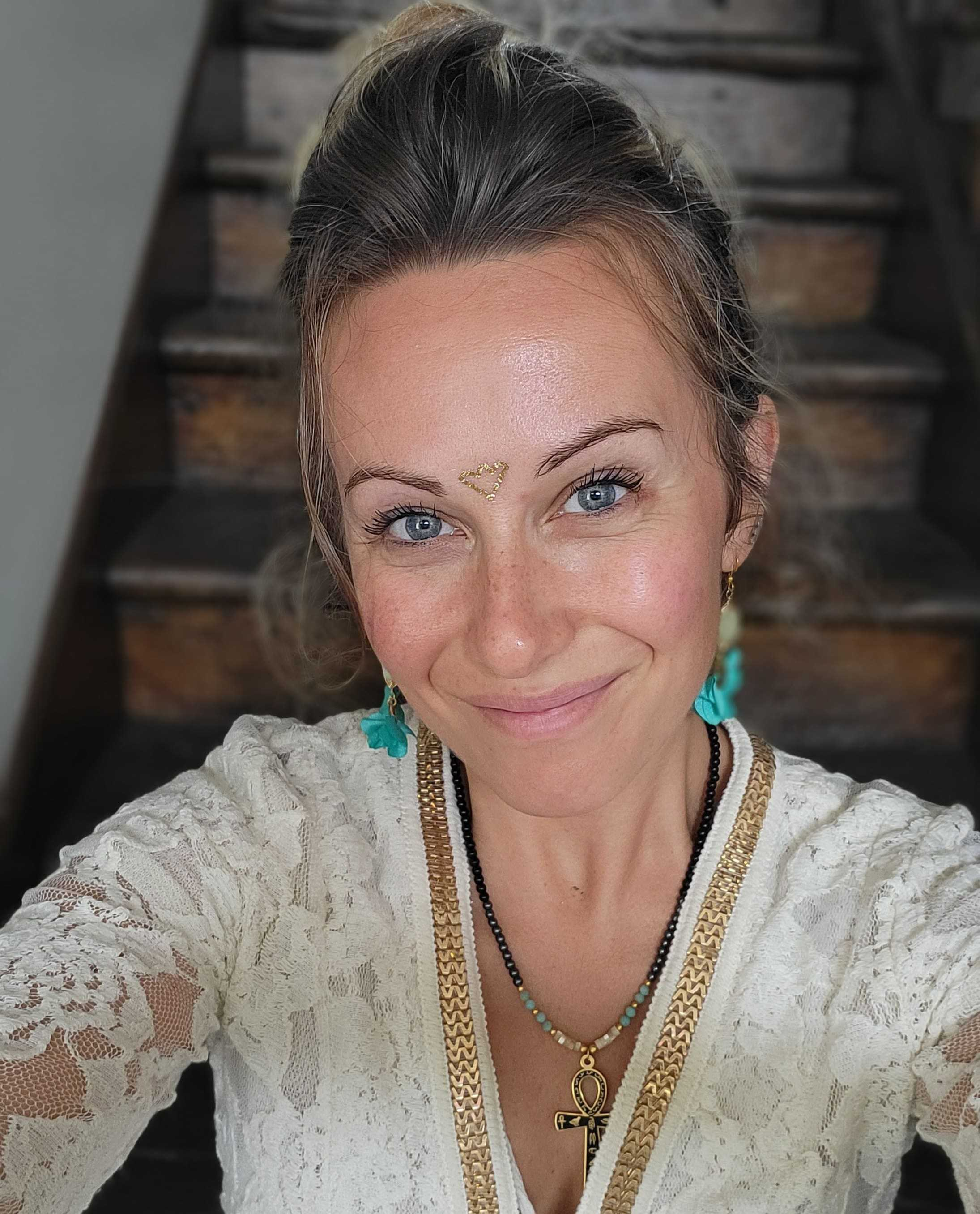 Une femme souriante avec des yeux bleus porte des bijoux dorés et un haut blanc en dentelle, avec un cœur doré dessinée sur son front.