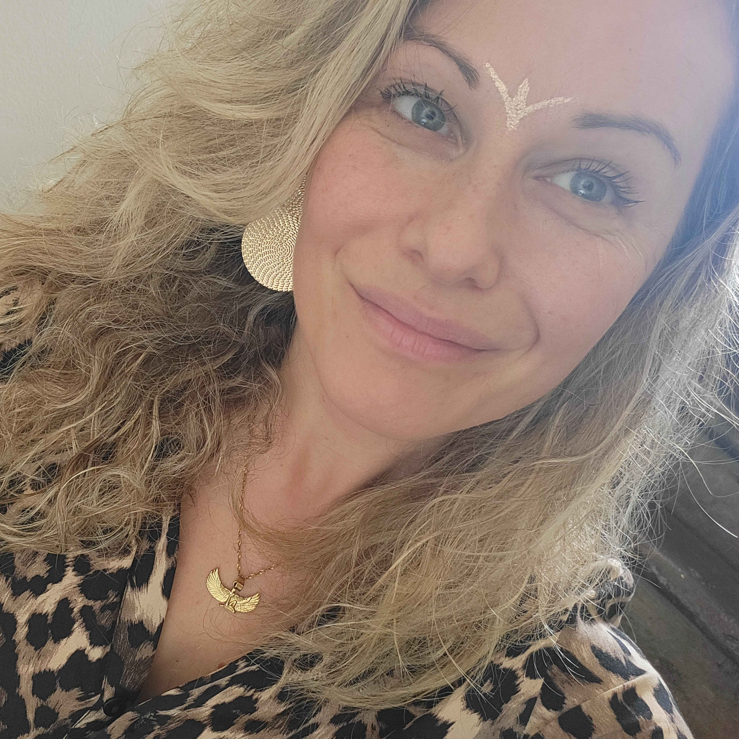 Une femme aux cheveux blonds ondulés, portant des bijoux dorés, un chemisier à motifs léopard et un collier avec un pendentif en forme d'oiseau, sourit à la caméra.