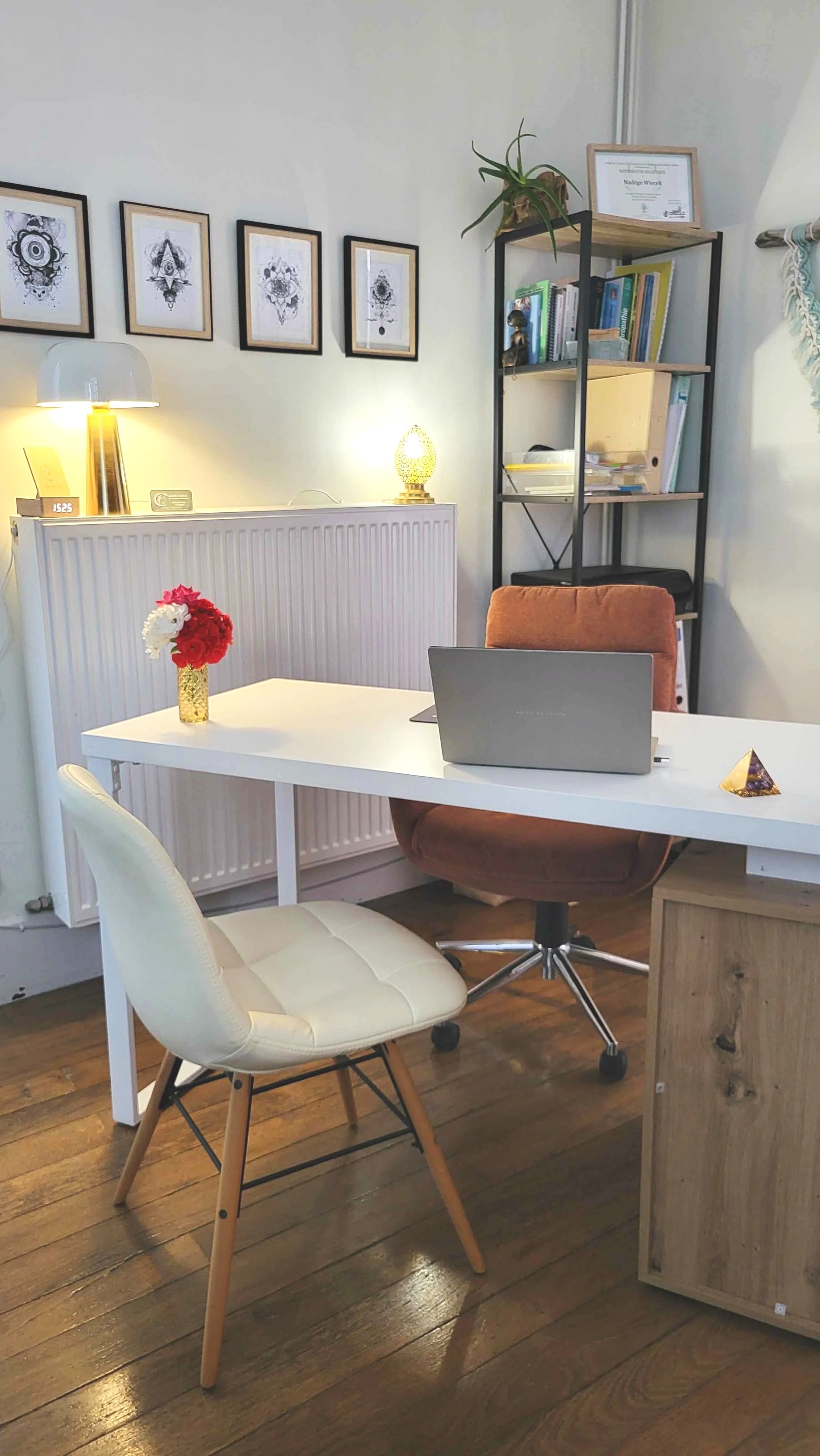 Un bureau avec une chaise blanche, une autre chaise orange, un ordinateur portable, une petite table avec un vase de fleurs rouges et blanches, une étagère avec des livres, une lampe et une décoration, et des œuvres d'art encadrées sur le mur.