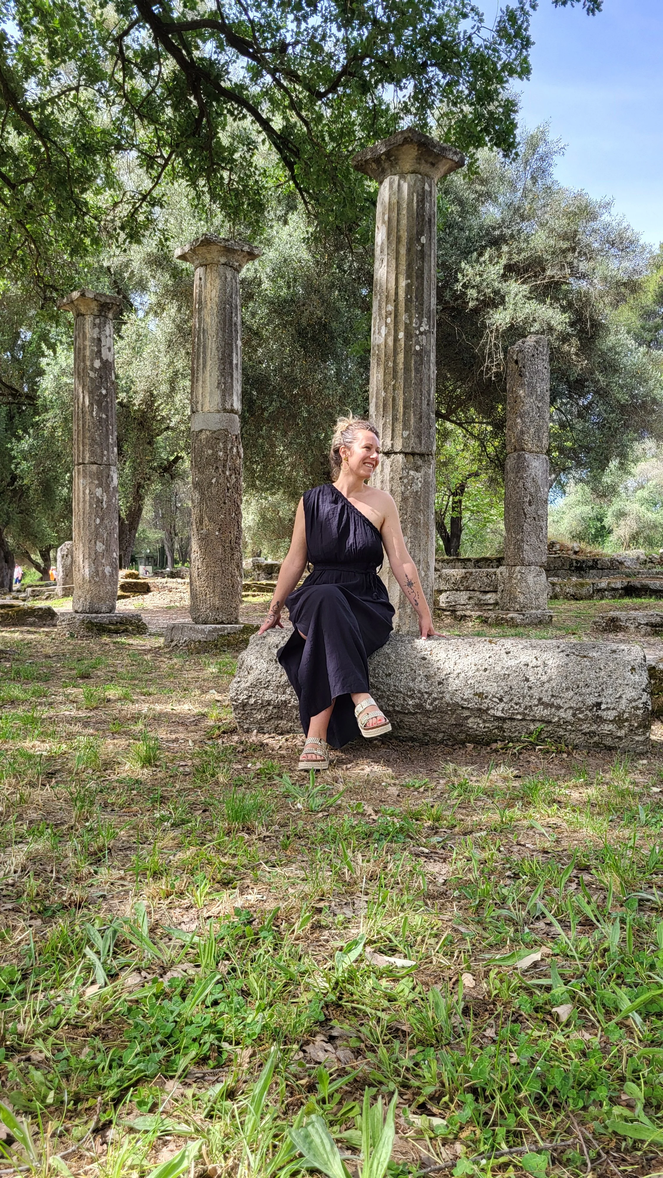 Une femme souriante assise sur une pierre dans un site archéologique avec des colonnes antiques en pierre, entourée d'arbres et de verdure.