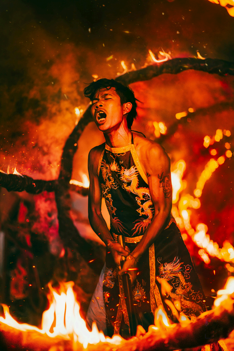 Une personne chante ou crie dans un environnement en feu, portant un vêtement traditionnel avec des motifs de dragon, entourée de flammes et de fumée.
