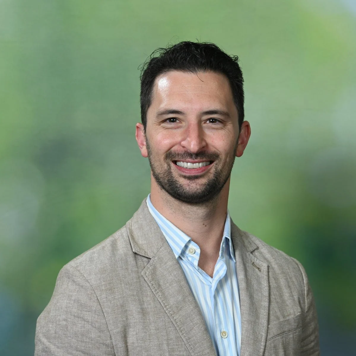 Aaron Gevers: A smiling man with dark hair and a beard, wearing a beige blazer and a light blue striped shirt, standing against a green background.
