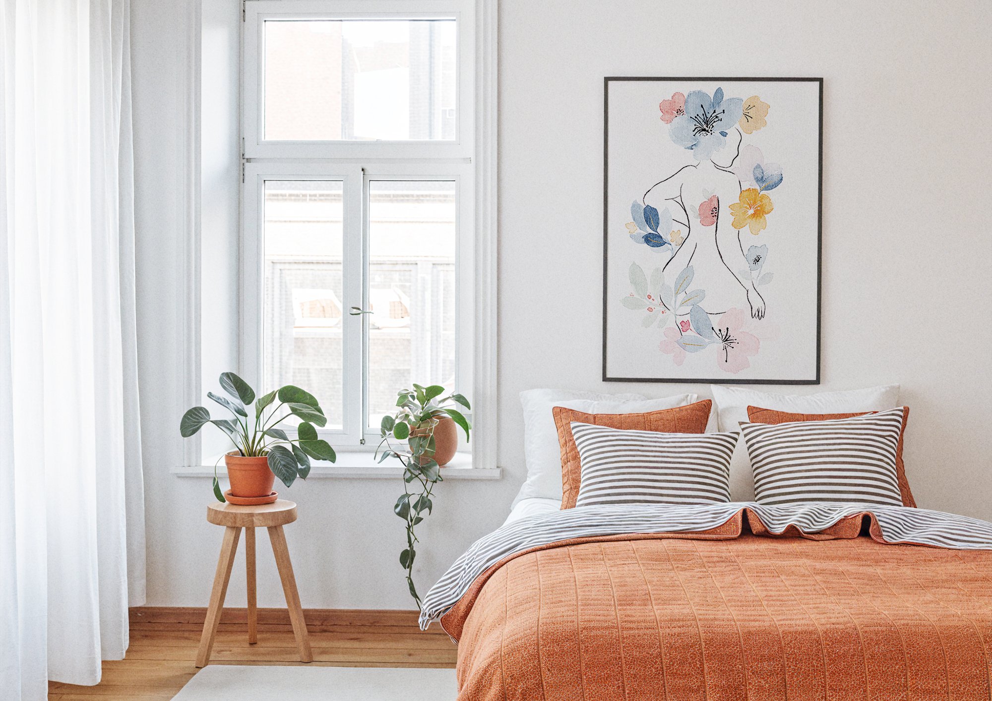 A cozy bedroom with a large window, white walls, and natural light. There are potted plants on a round wooden table, a bed with striped pillows and an orange duvet cover, and framed artwork depicting a line drawing of a woman surrounded by colorful f