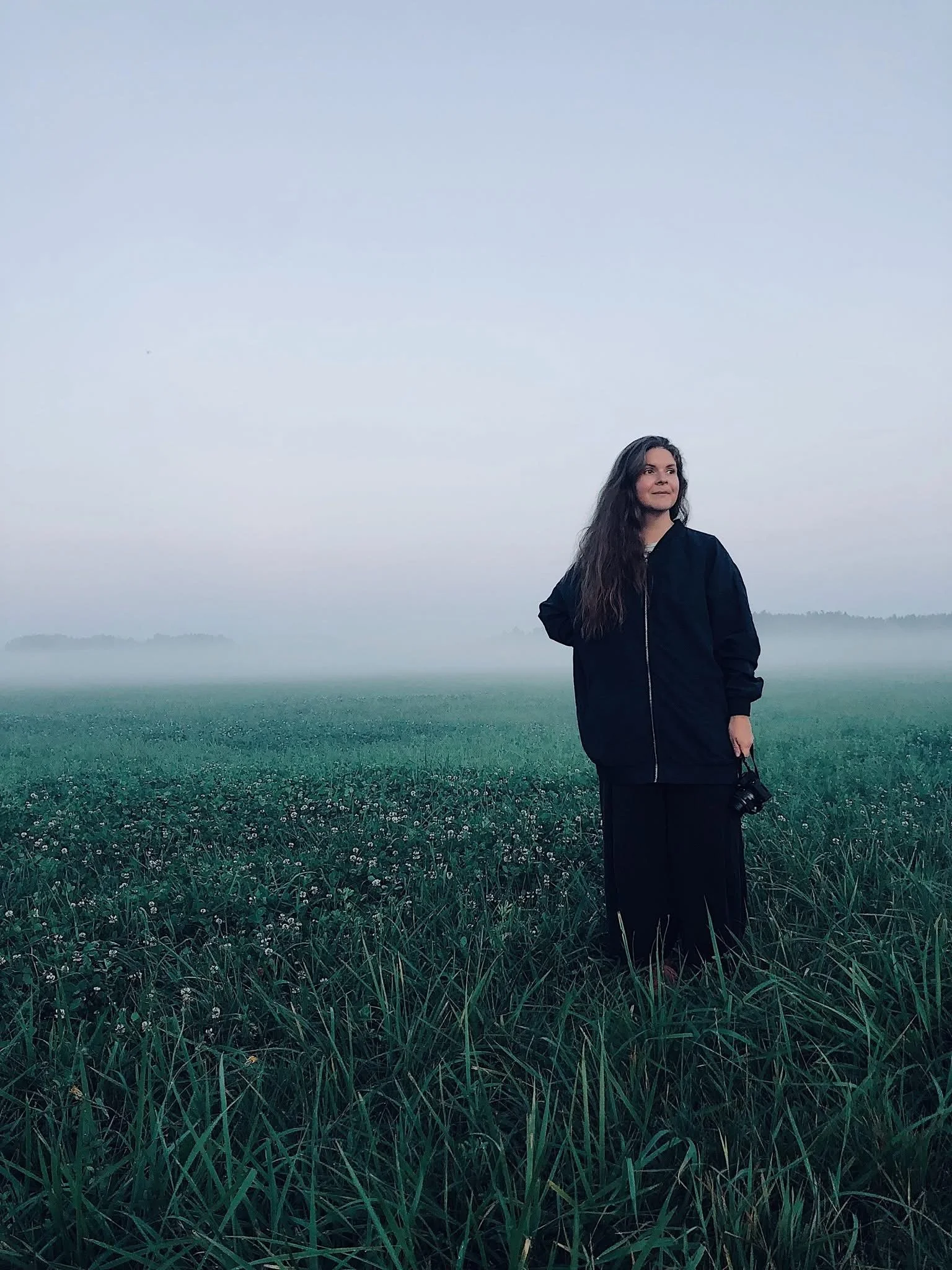 A woman with long dark hair wearing a black jacket and black pants stands in a misty field of tall green grass, holding a camera, with a light gray sky overhead.