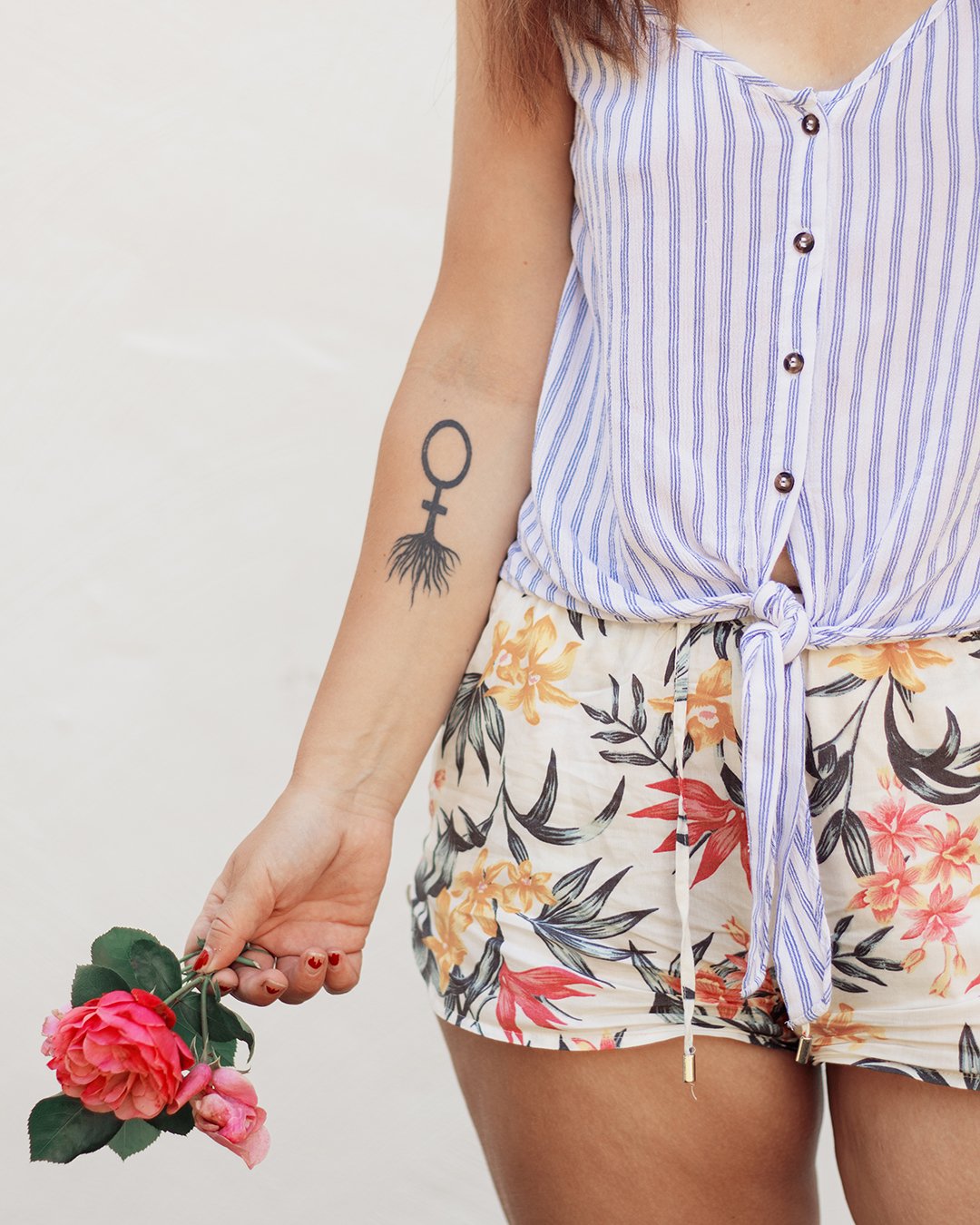 A woman holding a pink flower, showing a tattoo of a woman symbol with roots on her forearm. She is wearing a sleeveless blue and white striped tie-front top and floral shorts.