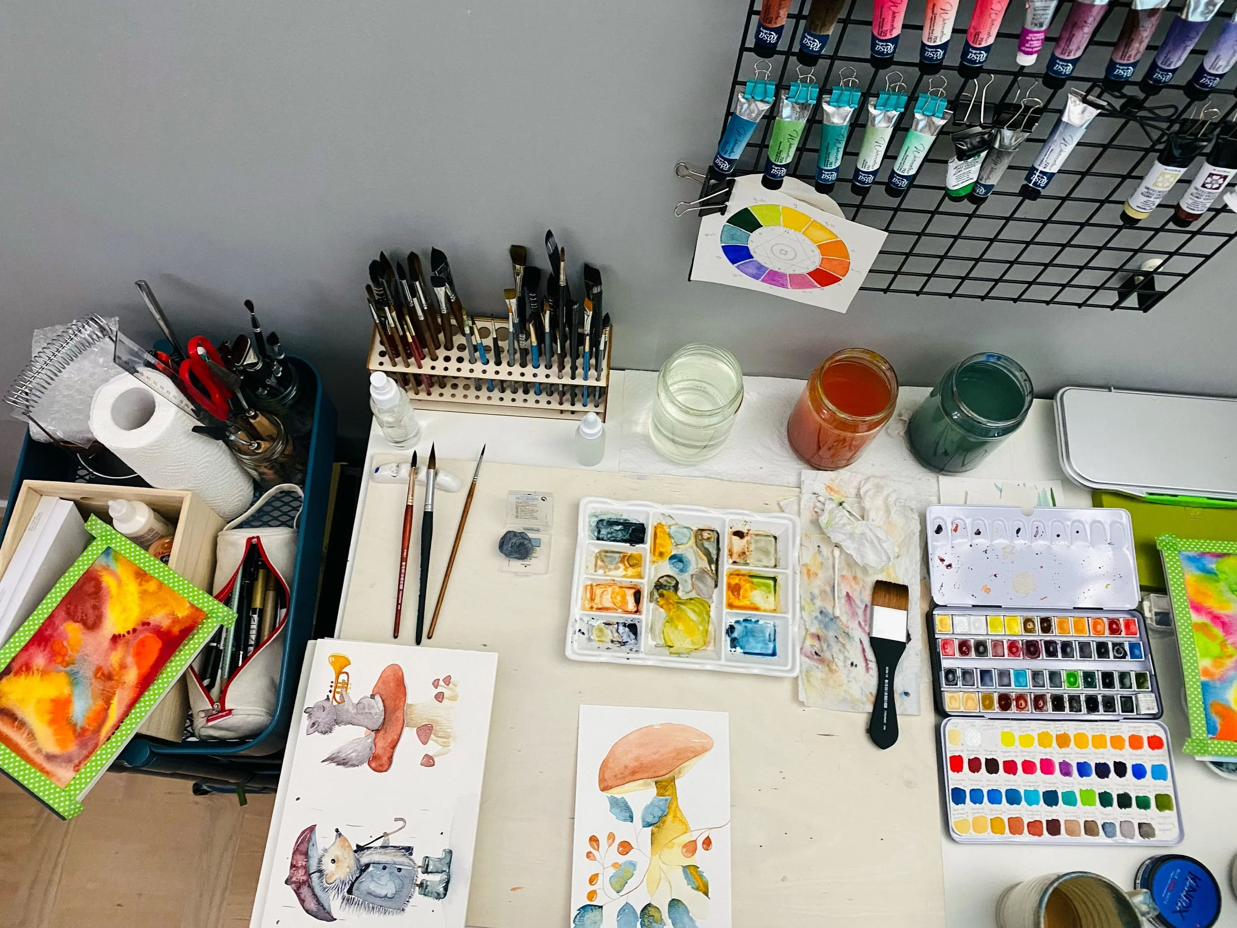 Art supplies including watercolor paints, brushes, paper with animal watercolor paintings, watercolor palette, jars of water, and paint tubes arranged on a workspace