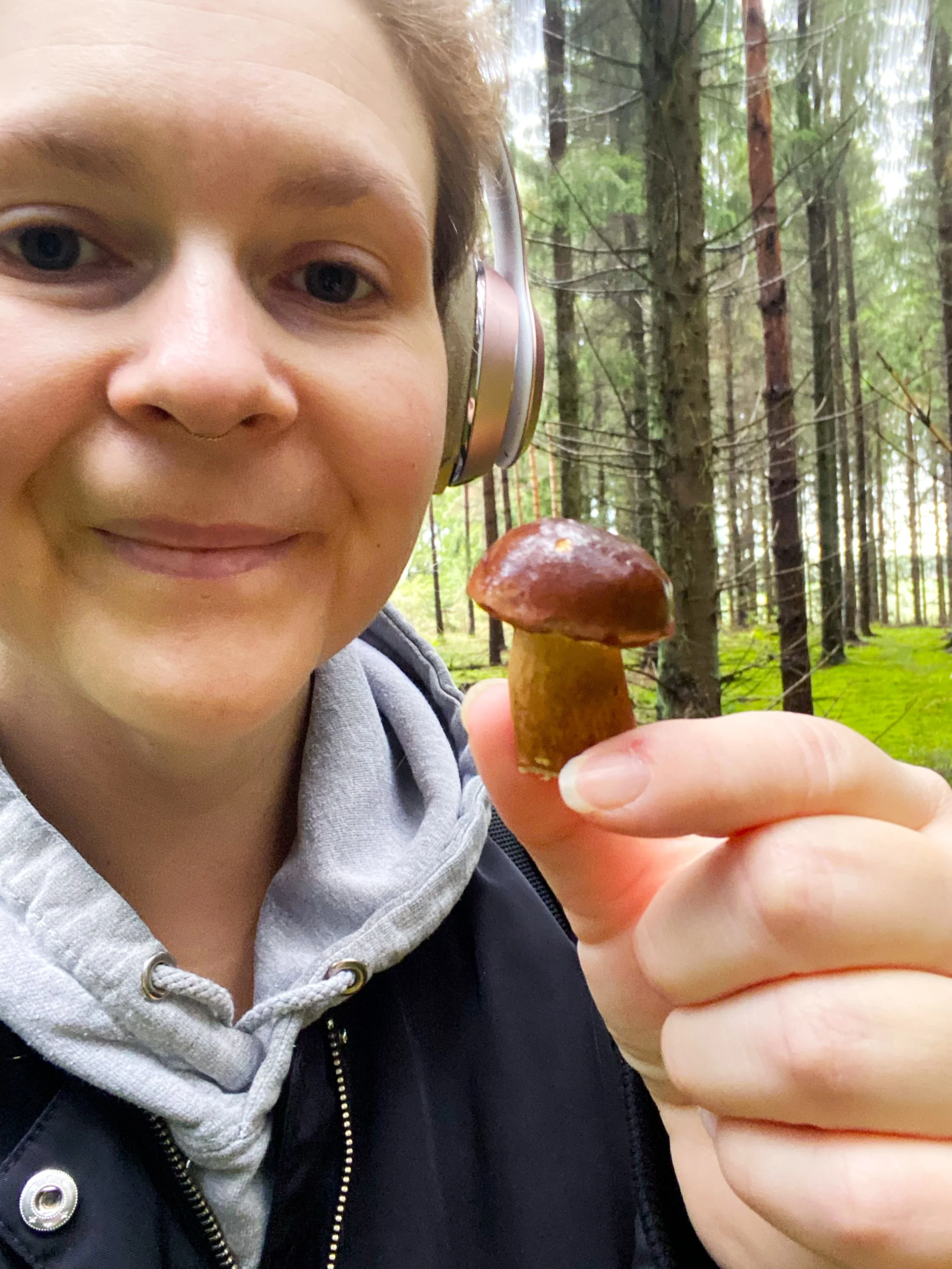 A woman with hair in a ponytail wearing headphones, smiling and holding a tiny mushroom in a forest with tall green trees.