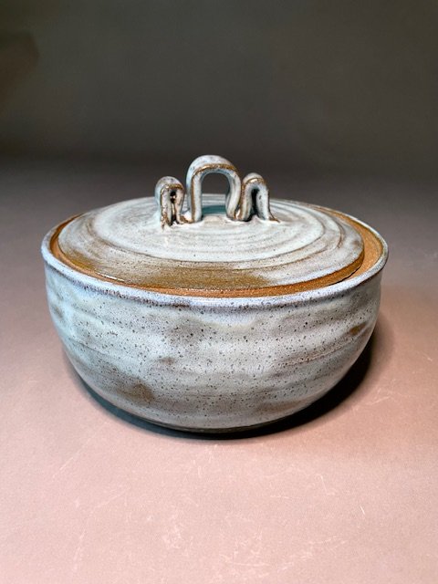 A round ceramic bowl with a lid, featuring a handle shaped with 3 curves, in a speckled light blue glaze on brown clay.