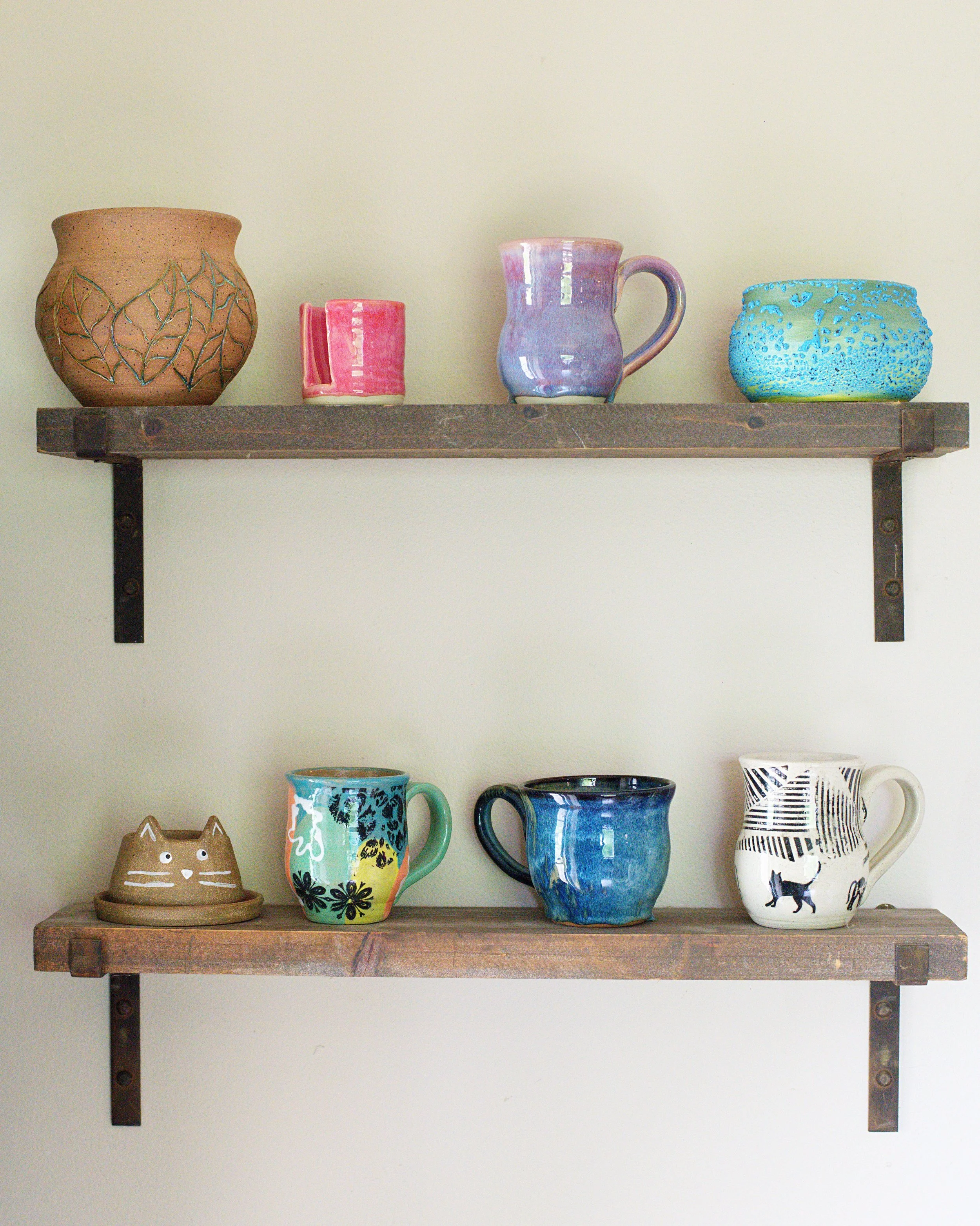 Display of various colorful ceramic mugs and containers on a two-tier wooden shelf against a beige wall.