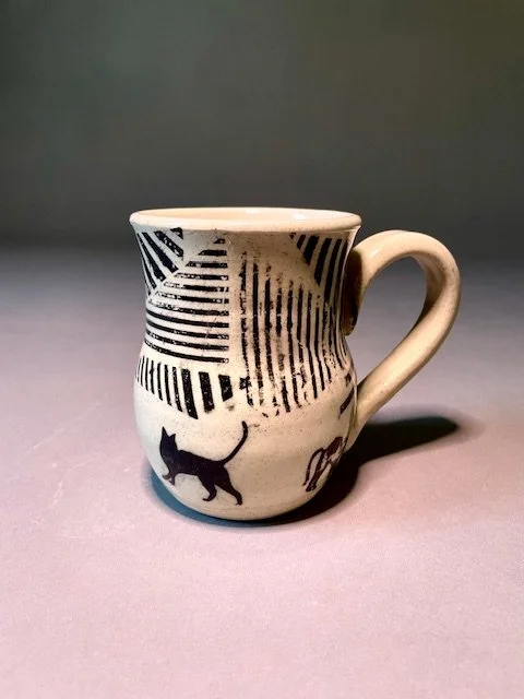 White ceramic mug with black abstract lines and a black silhouette of a cat on the front, placed on a gray surface with a gray background.