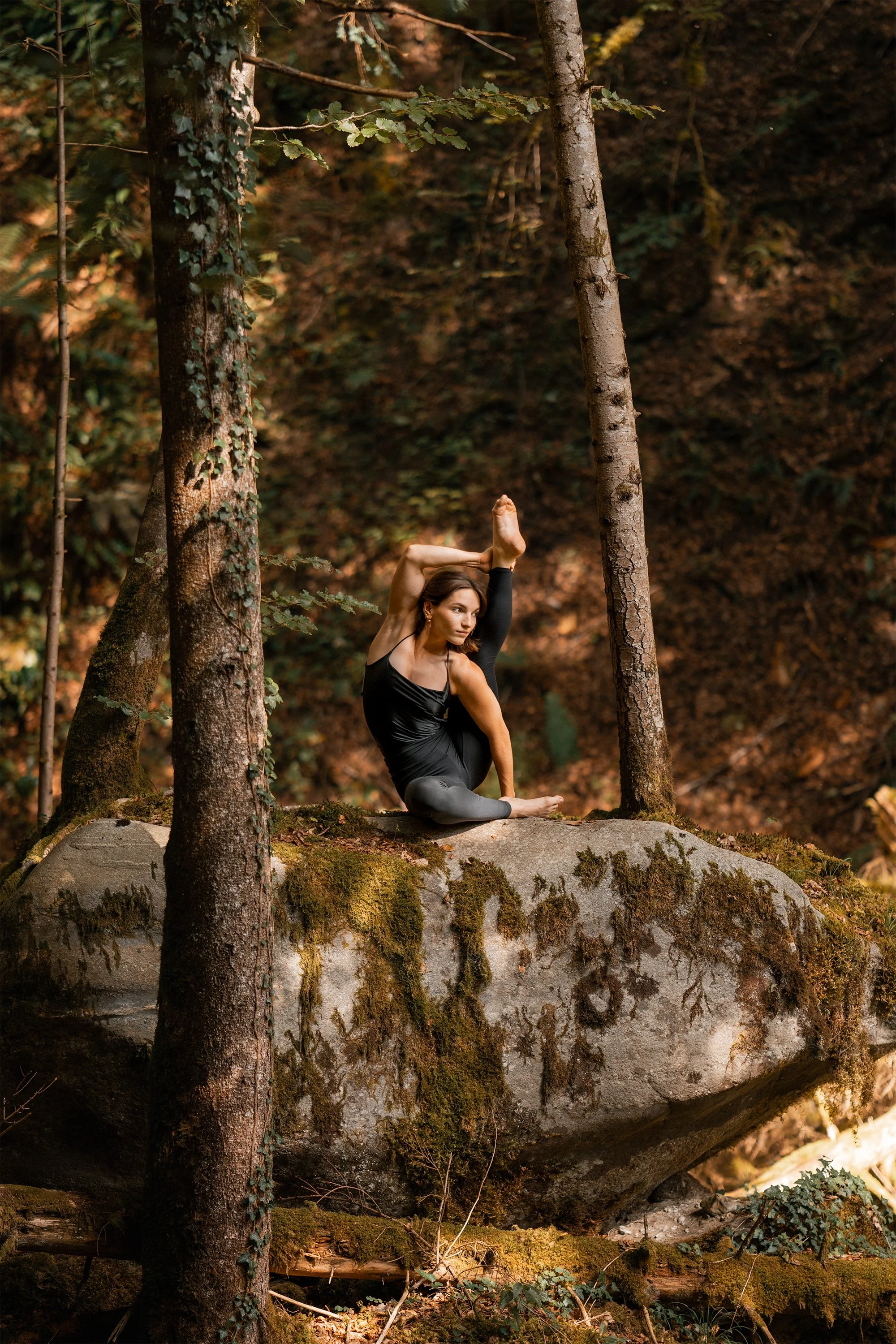 Une femme pratique le yoga dans la forêt sur une roche recouverte de mousse, entourée d'arbres et de feuillage automnal.